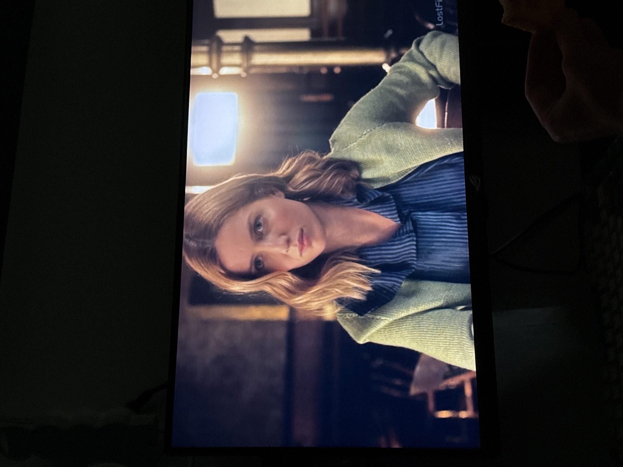 A woman with blonde wavy hair is seen on a television screen. She is wearing a blue striped shirt and a light green jacket. The background appears to be an interior location with a window and what seems to be metal fixtures. The city and location are unknown. A hand is partially visible in the upper right corner.