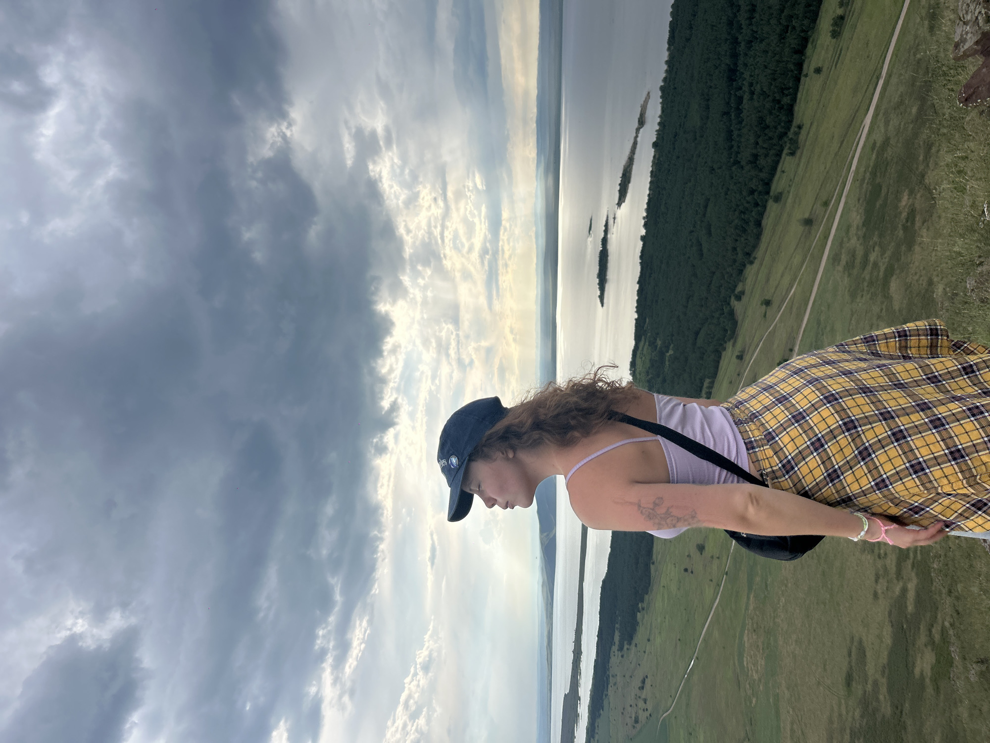 In the city of Nikolo-Petrovka, Russia, a young woman stands on a grassy hilltop, her back to the camera, looking out over a vast lake. The sky above is dramatic, with heavy, dark clouds interspersed with shafts of sunlight. The woman is wearing a lavender tank top, a yellow and purple plaid skirt, and a dark baseball cap. She has curly brown hair and a tattoo on her left arm. A dark shoulder bag is slung across her chest. The landscape features rolling green hills dotted with trees, leading down to the expansive blue water. Islands can be seen in the distance on the lake, and a winding dirt road is visible on the hillside.