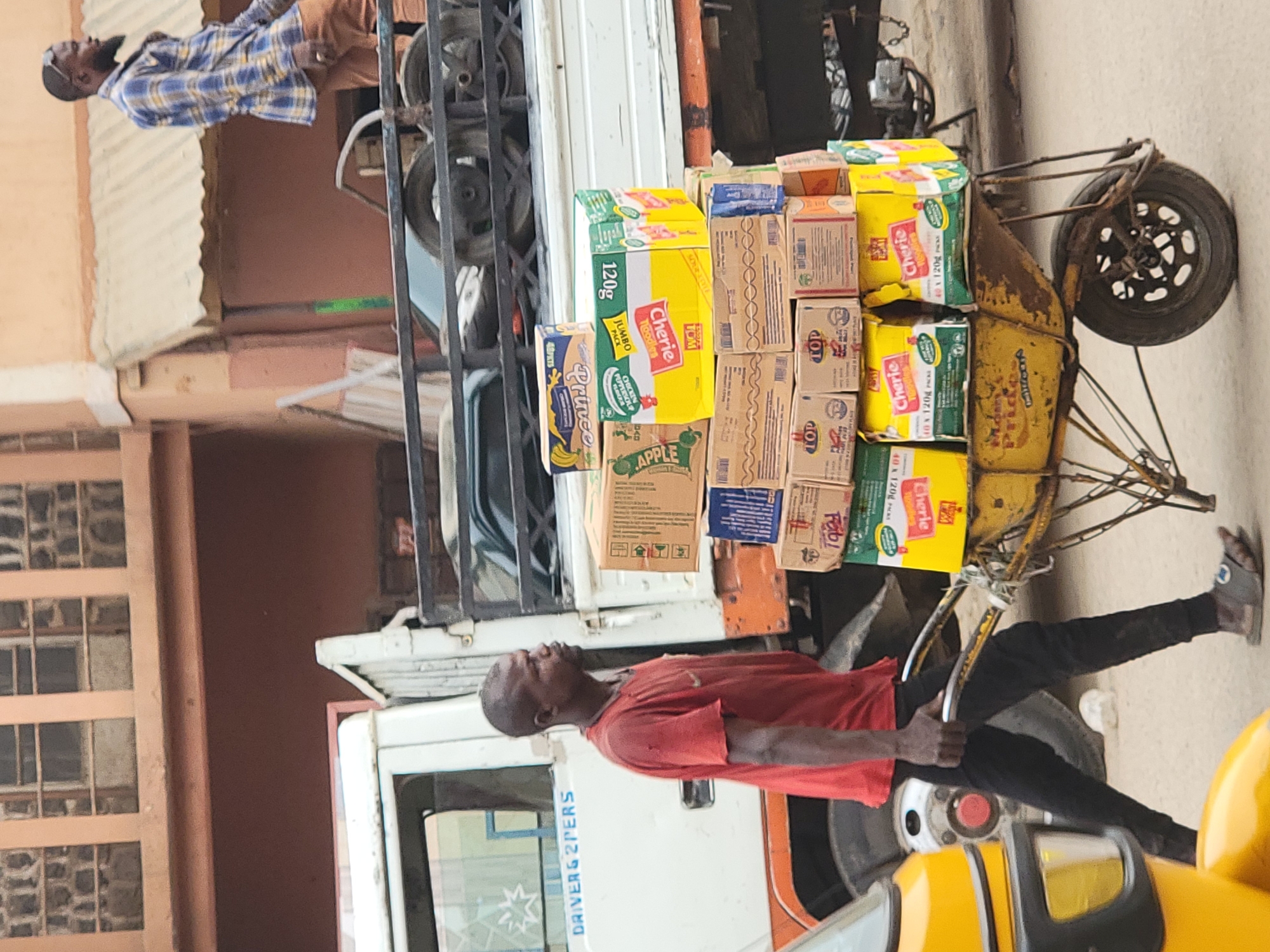 This image, taken in Maiburiji, Nigeria, captures a daytime street scene featuring two men and a heavily loaded handcart.
In the foreground, a man wearing a red t-shirt and dark trousers is actively pushing or maneuvering a yellow, well-worn handcart. The cart is piled high with numerous cardboard boxes containing various products. His focus appears to be on the task of transporting these goods.
The boxes on the handcart are clearly visible and bear several brand names and details. Prominent text includes "Cherie noodles JUMBO PACK," with associated weight "120g," and multiple boxes simply marked "Cherie." Other discernible labels include "TOP," "Fab," "Prince" (with a banana graphic), and "APPLE" (with an apple graphic). Some boxes also indicate quantities like "20 x 120g" and "40 x 120g."
Behind the man with the handcart, a white truck or van is partially visible. On its side, the text "DRIVER & 2PERS" can be read, likely indicating its passenger capacity. Another man, dressed in a blue and yellow plaid shirt and brown trousers, stands on what appears to be the back or an adjacent platform of this vehicle, looking towards the left side of the frame.
The background shows buildings with corrugated metal roofing and window grates, suggesting an urban or semi-urban environment. The scene is brightly lit, indicating clear daytime weather. The image depicts a common method of goods transportation in the region, highlighting daily commerce and activity.
