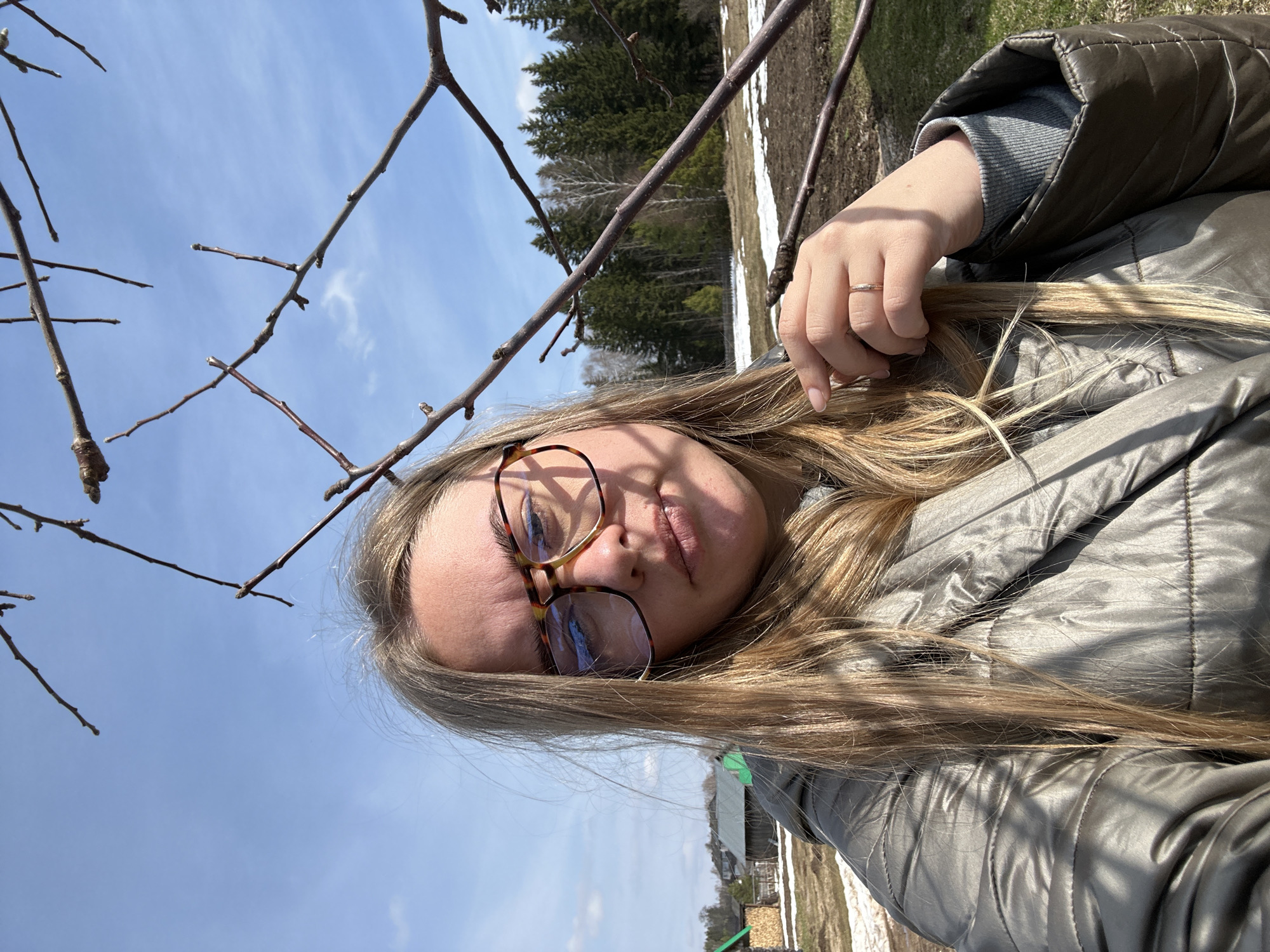 The image shows a selfie of a person with long blonde hair and eyeglasses, wearing a gray jacket. The person is holding a strand of their hair with their right hand, which has a ring on the ring finger. In the background, there is a blue sky with bare tree branches in the foreground. Behind the branches, there is a view of a forest and a field with patches of snow. There also appears to be some kind of building. The location is in the city of Уральское, Russia.