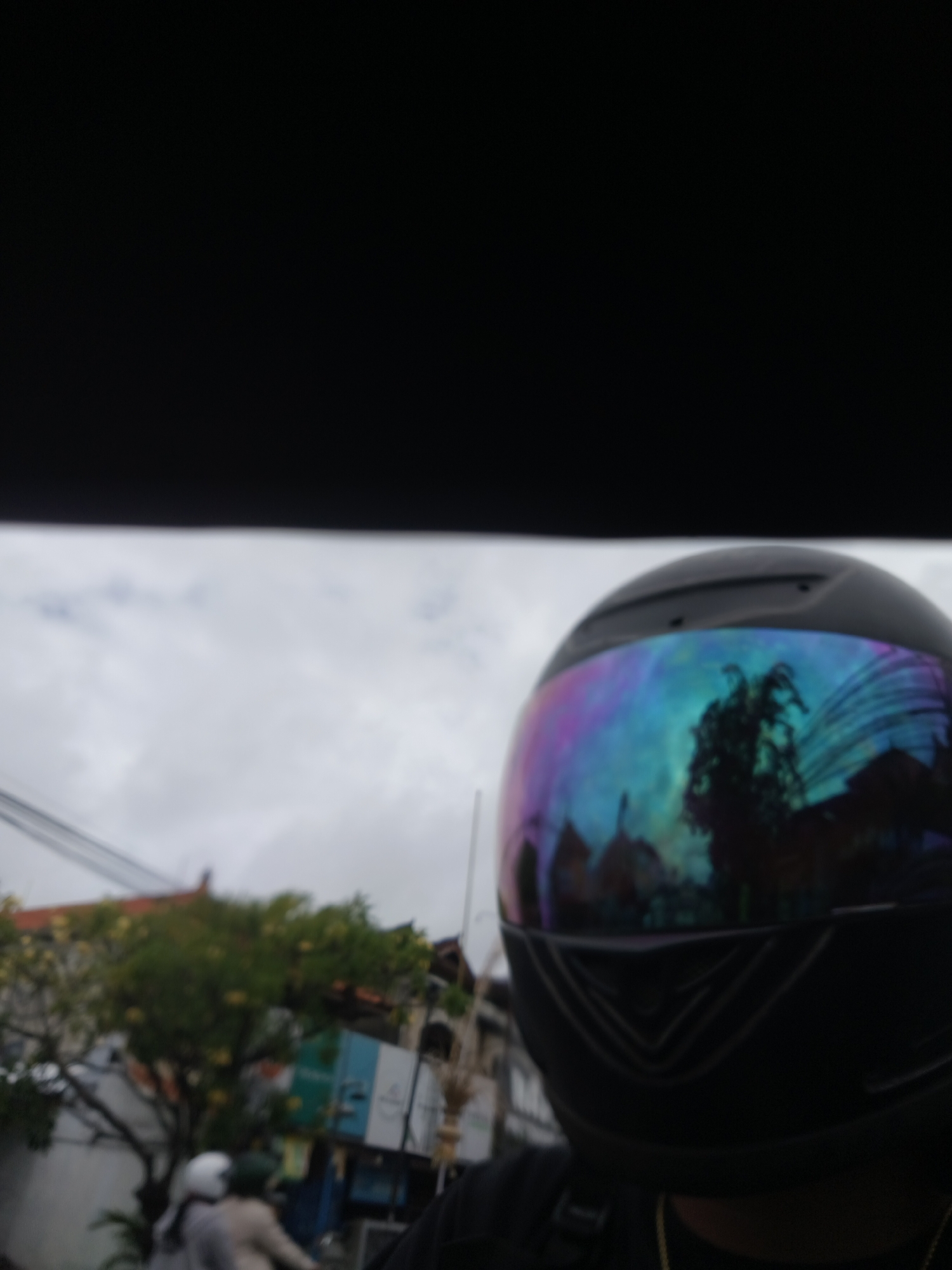 A person wearing a black helmet with a multicolored, reflective visor is in the foreground of the image. The visor reflects trees and buildings, suggesting an outdoor environment. In the background, there are trees with yellow flowers, buildings, and electrical wires, indicating an urban setting. Two people on a motorcycle are partially visible in the lower left of the frame, also suggesting activity within a city. The sky is overcast. The scene appears to be a street in Denpasar, Indonesia.