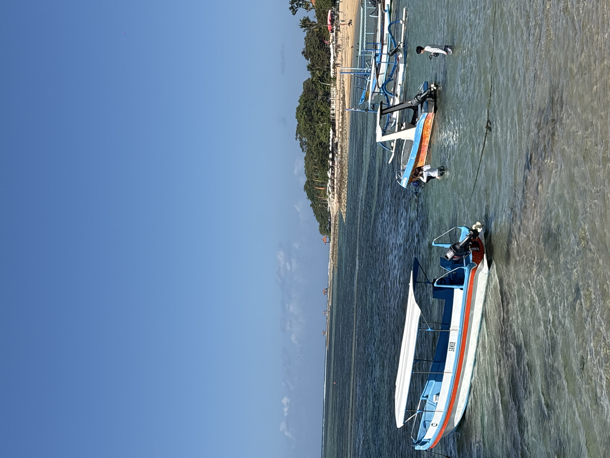 The image depicts a tropical beach scene in Denpasar, Indonesia, under a clear blue sky. In the foreground, several boats are moored in shallow, clear turquoise water. The closest boat, a long, narrow vessel with a white canopy, has "SANUR" written on its side. Further out, two other boats, one orange and white and another larger white boat with blue accents, are visible. Two individuals, a child and an adult, are in the water near the orange and white boat, one appearing to interact with it.
In the middle ground, a sandy beach is lined with numerous white umbrellas, suggesting a resort or tourist area. Behind the beach, lush green trees form a dense backdrop. Several small, open-air gazebos are situated on the beach and in the water, some with thatched roofs. The water extends to the horizon where a faint line of clouds is present. A small pink object, possibly a balloon or kite, is visible high in the sky. The scene captures a serene day at a popular Indonesian beach destination.