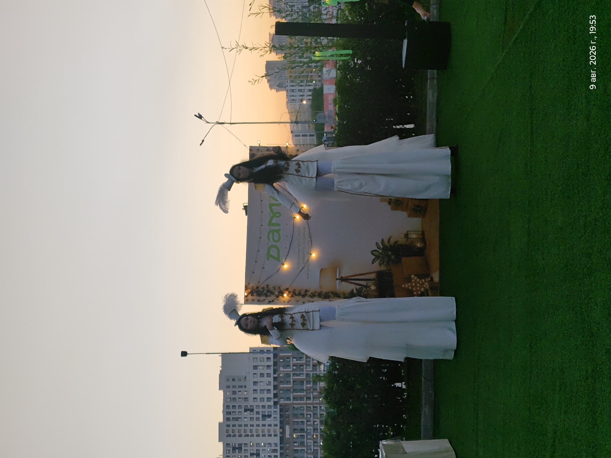 Two performers on stilts, dressed in elaborate white costumes with feathered hats, stand in front of a backdrop with the logo "Dama" and string lights. They are outdoors on a patch of artificial grass, with buildings visible in the background. The sky is a soft gradient of orange and light blue, suggesting either dawn or dusk. The scene appears to be an outdoor event or performance.
