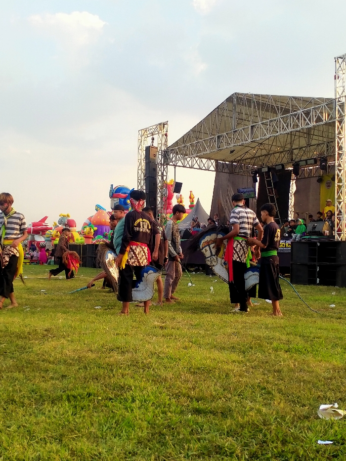 The image displays an outdoor cultural event in a grassy field within the city of Glonggong, Indonesia, under a bright, partially cloudy sky. In the foreground and midground, a group of approximately 10-15 individuals, predominantly young males, are present. Several are dressed in dark pants with patterned batik fabric wrapped around their waists and some are barefoot. They are associated with a Kuda Lumping (Ebeg) performance, holding or wearing stylized horse puppets made of white material with blue and black accents. One individual in the mid-left is positioned forward, holding a horse puppet. In the background, a large, silver metal truss stage with a flat roof and prominent speaker stacks is erected. To the left of the stage, multiple large, colorful inflatable structures, including forms resembling a robot, a rocket, and abstract shapes, are visible. White litter is scattered across the grass in the foreground.