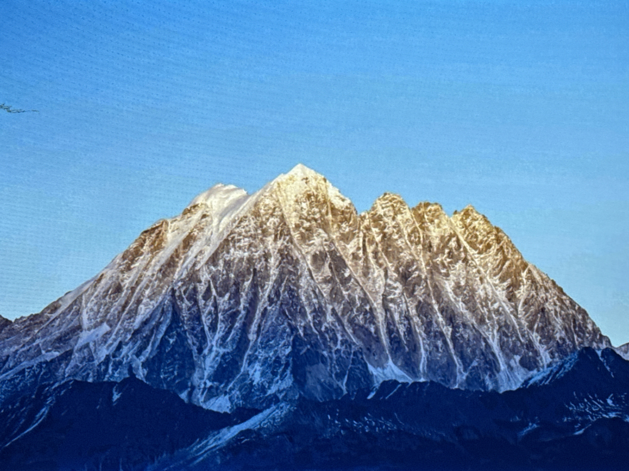 This image shows a vast mountain range under a clear, bright blue sky. The mountains are snow-capped and rugged, with sharp peaks and deep valleys. The sunlight highlights the snow-covered ridges, creating a dramatic contrast with the shadowed slopes. The lower parts of the mountains are dark and appear to be covered in dense vegetation or rock. There are no people or man-made structures visible in the image. The scene is one of natural grandeur and remoteness. The provided location information "Жәпек батыр ауылы, Kazakhstan" does not appear to be directly relevant to the visual content of the image as there are no urban or rural elements identifiable.