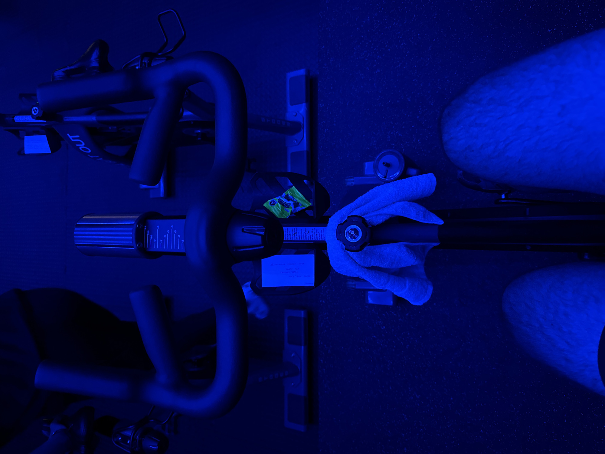 An overhead shot captures an indoor cycling (spinning) setup in a gym or studio in San Bernardino Tlaxcalancingo, Mexico, under pervasive blue lighting.
**Subjects:** The scene features an exercise bike and parts of a person. The person's lower legs and feet are visible on the right, clad in what appears to be furry slippers or heavily textured socks/footwear.
**Objects:** The exercise bike's handlebars, frame, and console dominate the left and center of the image. A resistance control knob, labeled "STOP," is visible on the bike's frame, with a small white towel draped over it. Adjacent to the towel, a water bottle rests upright. A small, colorful wrapper (possibly for candy or a snack) is placed on the bike's console. The console itself displays numerical markings, likely indicating resistance or intensity levels. A handwritten sticky note is attached to the bike frame.
**Activity or event taking place:** The presence of the exercise bike, towel, water bottle, and a note detailing workout statistics strongly suggests the person has either just completed or is about to start an indoor cycling workout. The top-down perspective places the viewer directly over the equipment and user.
**Notable details:** The entire scene is bathed in a deep blue light, creating an atmospheric, perhaps low-light, environment typical of spin classes. The floor has a speckled, dark texture. The sticky note provides specific, legible workout data: "SESSION DONE: 100", "DATE: MAY", "TIME: 18:30", and "DISTANCE: 25.0".
**Visible text:**
* "OUT" (partially visible on the bike frame/handlebar)
* "STOP" (on the resistance control knob)
* On the sticky note: "SESSION DONE: 100", "DATE: MAY", "TIME: 18:30", "DISTANCE: 25.0"
* Numerical markings on the bike console (e.g., -10, -5, 0, 5, 10).