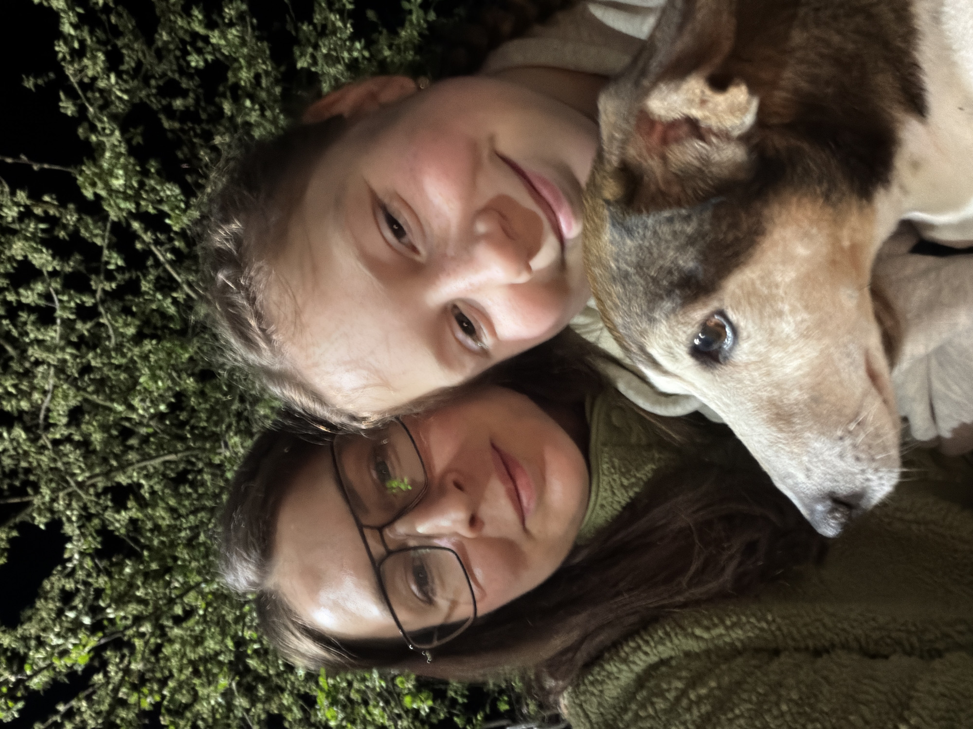 A low-angle, close-up selfie shows two people and a dog. The person on the lower portion of the frame has shoulder-length brown hair and wears glasses. The second person, above the first, is younger and has lighter hair. A brown and white dog is between them. The background is dark with green foliage, potentially bushes. The image appears to be taken outdoors at night. The location is unspecified, so the city of Markovo, Russia, cannot be confirmed or denied as the setting.