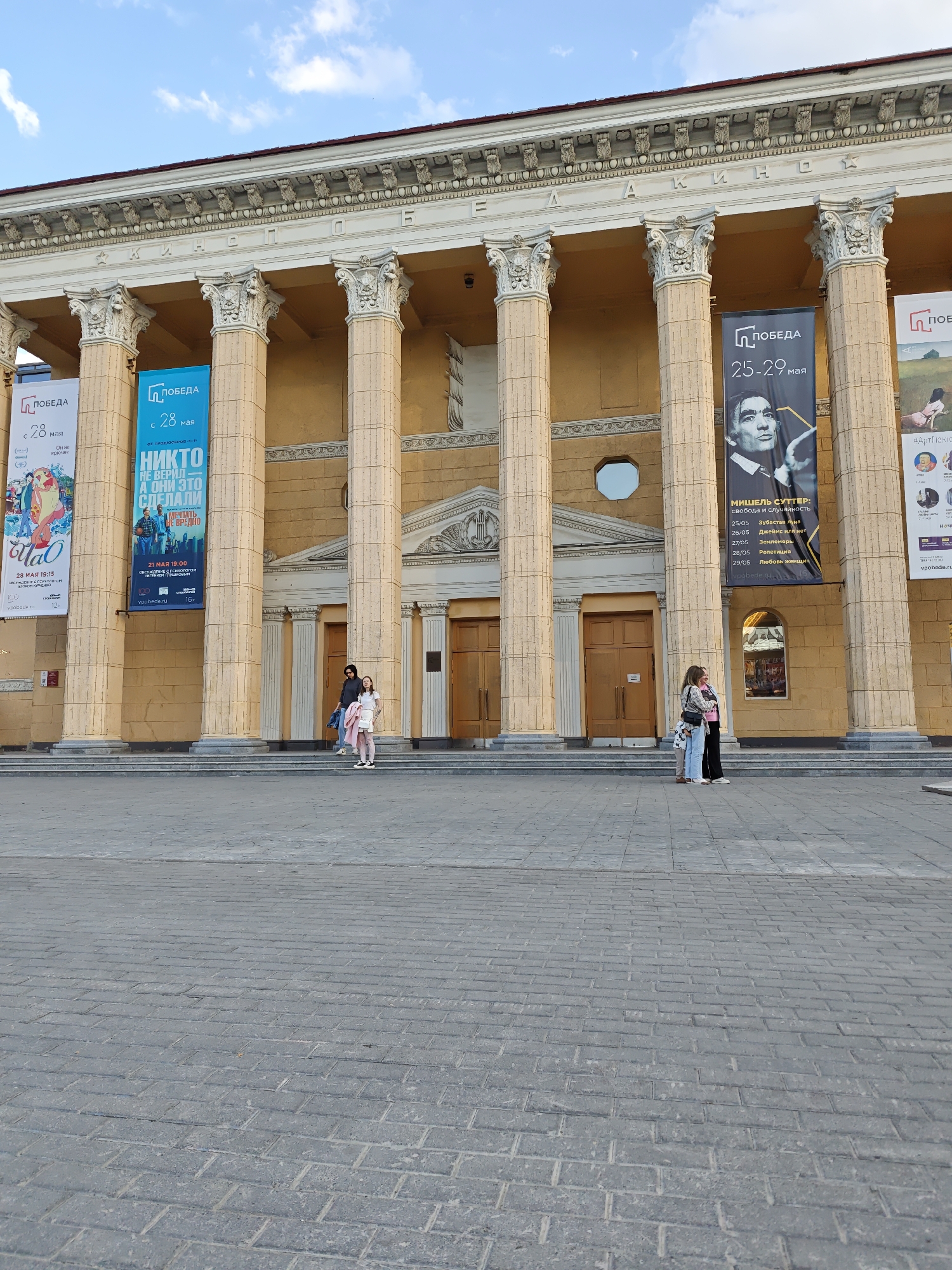 The image shows the facade of the "Pobeda" Cinema in Novosibirsk, Russia. The building features a classical architectural style with a row of tall columns. Banners advertising upcoming events hang between the columns. Several people are standing on the steps leading up to the cinema. The foreground shows a paved area. The sky is visible above the building.