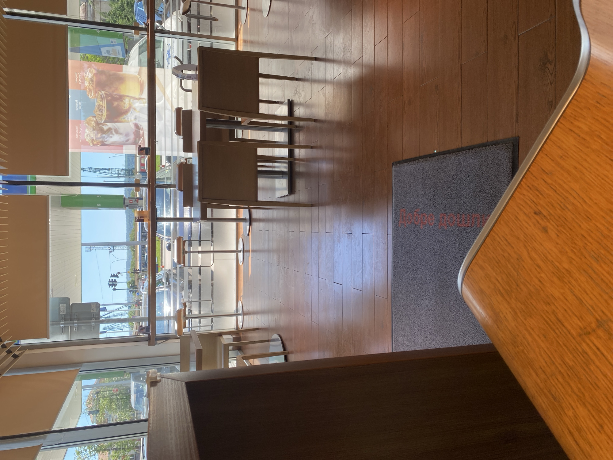 The image depicts the interior of a cafe, possibly in Varna, Bulgaria, judging by the Cyrillic lettering on the entrance mat ("Добре дошли," meaning "Welcome"). The scene is framed by a section of a wooden countertop in the foreground. The cafe features seating arrangements including tables and chairs, visible through a large glass window. Outside the window, there are views of the urban landscape, including a gas station, traffic lights, and other street furniture. Light filters through the blinds, suggesting it is daytime. The interior design includes wooden flooring. There are no people immediately visible in the frame.