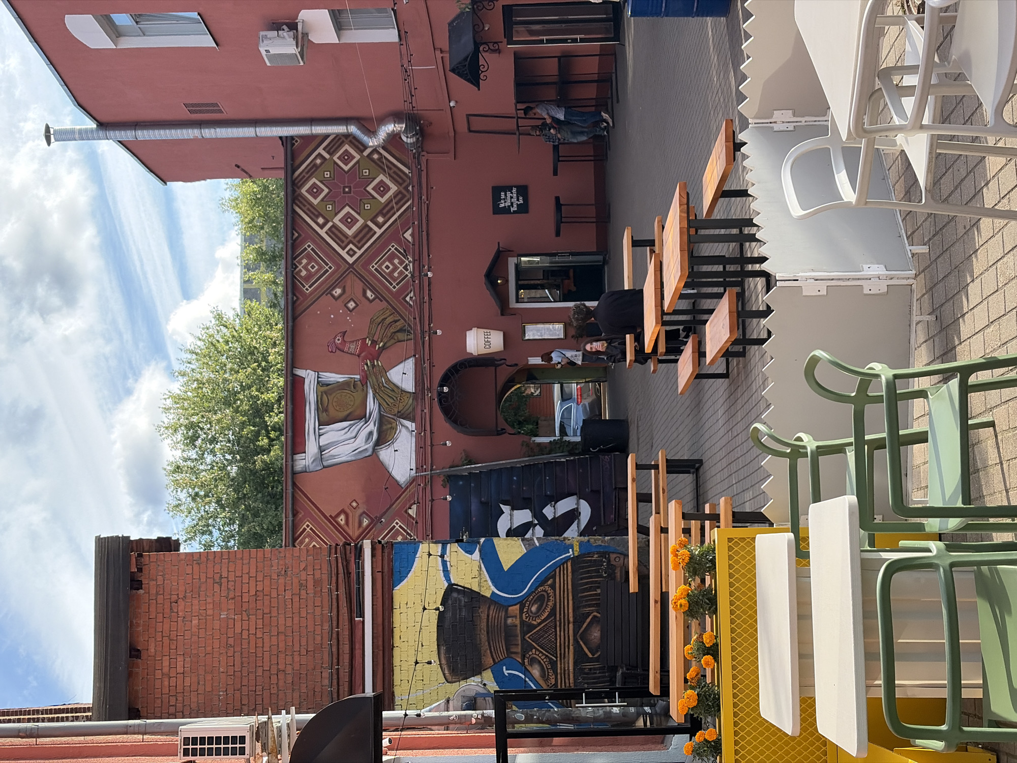 The image shows an outdoor courtyard area of a coffee shop located in Minsk, Belarus. The courtyard features several picnic tables with wooden benches, interspersed with white tables and green chairs. A yellow planter box with orange flowers is visible in the foreground. The walls of the surrounding buildings are painted in a reddish-brown hue and adorned with murals. One mural depicts a stylized human face with a headscarf and a bird, while another features a large, decorative vase. A set of dark stairs leads up to an arched entryway of a building that houses the coffee shop, indicated by a white "COFFEE" sign. Several people are scattered throughout the courtyard, some sitting at tables, others standing in small groups. The ground is paved with light-colored bricks. The sky above is partly cloudy.