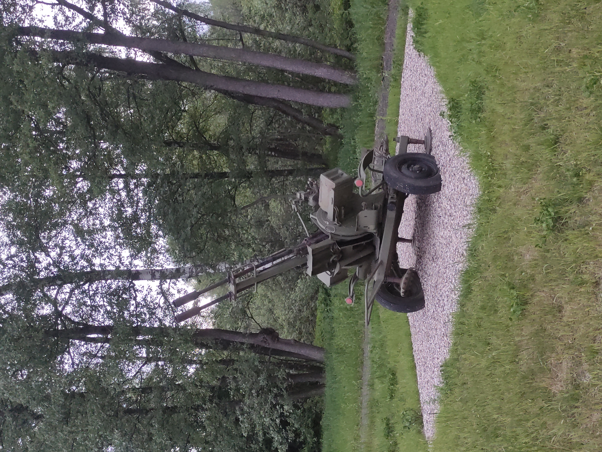 A static, broad shot shows a green artillery piece, possibly a ZU-23-2 anti-aircraft gun, positioned on a light gray gravel path. The path divides a wooded area to the left, consisting of trees with dense green foliage, from a grassy field to the right. The scene appears to be outdoors, possibly in a park or memorial area. The location is identified as Shevelkino, Russia. There are no people visible in the image.