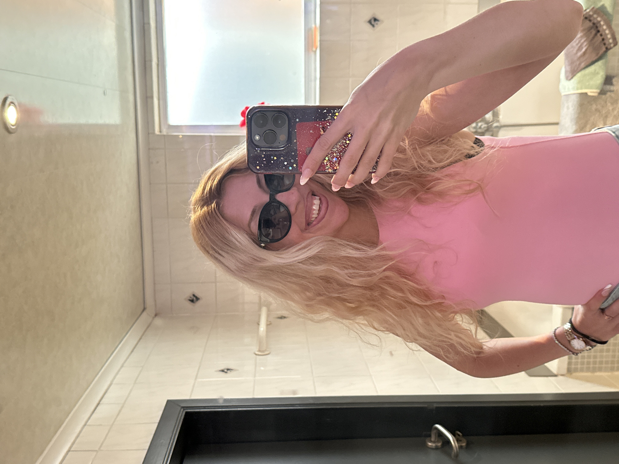 A woman with long blonde wavy hair, wearing sunglasses, a pink top, and two bracelets, is taking a mirror selfie in a bathroom. She is smiling and sticking her tongue out. The bathroom has tiled walls, a metal door handle, and a window. There is a phone in her hand with a glittery case.