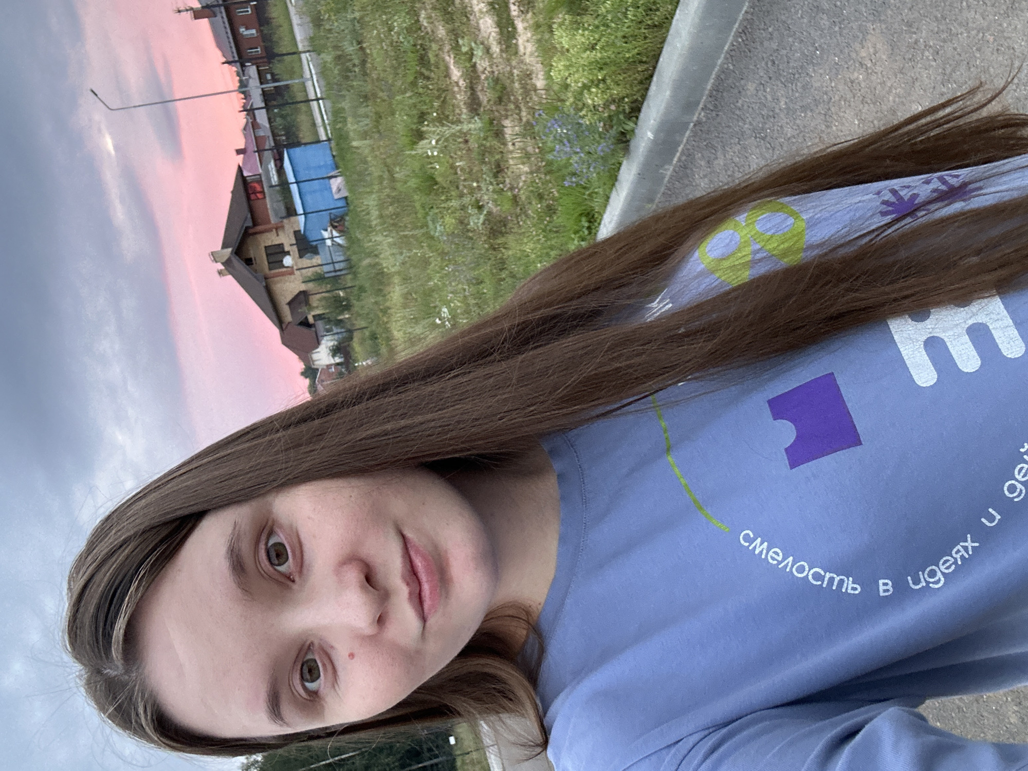 The original image is a selfie of a young woman with long, brown hair and fair skin. She is wearing a light blue long-sleeved t-shirt with some graphics and text on it. The text on the shirt is in Russian and appears to be in a circular pattern. There are also graphics of a purple puzzle piece and some green shapes on the shirt.
The woman is standing outdoors, with a blurred background. The background shows a street or road in the foreground, with a grassy area and some vegetation to the right. In the middle ground, there are houses with red roofs and a blue shed. The sky in the background is cloudy with hints of pink, suggesting it is either sunrise or sunset.
The location is identified as Noviy, Russia. The overall scene is casual and appears to be taken in a suburban or rural setting.