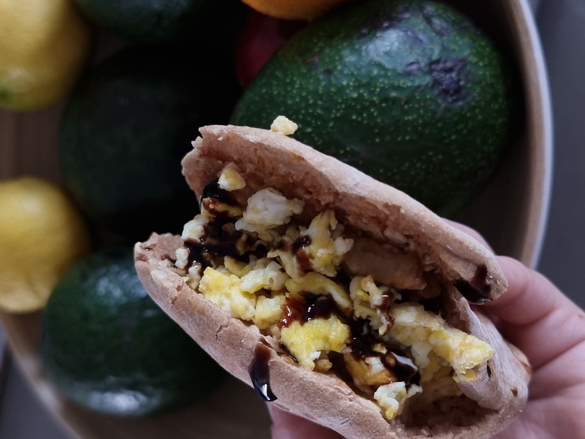 A person holds a pita bread sandwich filled with scrambled eggs and a dark sauce. Behind the sandwich, out of focus, are avocados and lemons in a wooden bowl. The location is Ramat Gan, Israel.