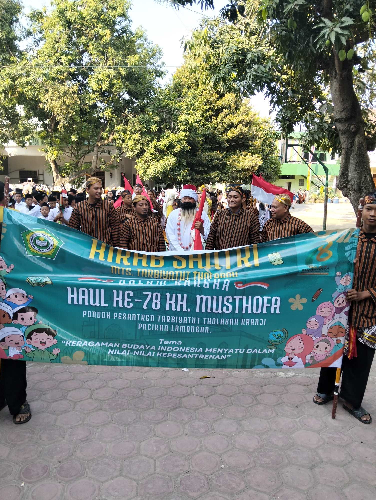 In Kranji, Indonesia, a group of people are participating in a parade. The central focus is a large green banner held by several individuals. The banner features text in Indonesian, indicating it's for the "Haul Ke-78 KH. Musthofa" and a theme related to Indonesian cultural diversity and pesantren values. Some people in the foreground are wearing traditional Indonesian clothing, including striped shirts and head coverings. Behind them, a crowd of younger people, likely students, are also visible, some holding Indonesian flags. The event appears to be taking place outdoors, with trees and buildings in the background. The ground is paved with hexagonal tiles.