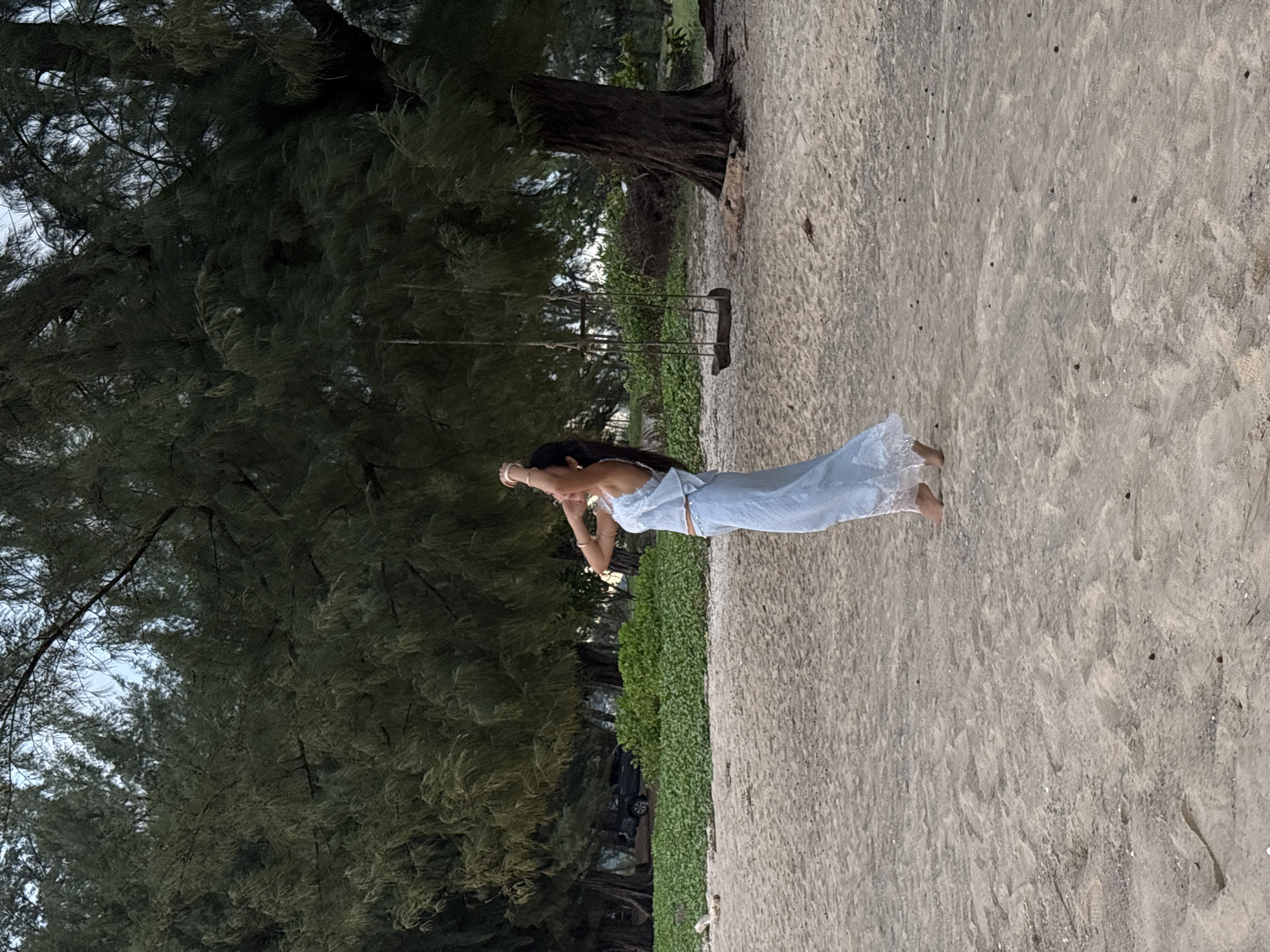 An outdoor, eye-level, full shot captures a woman from the knees up, standing on a sandy beach, facing right. She is wearing a light blue two-piece outfit adorned with white lace trim along the hem of her top and the bottom of her pants. Her arms are raised above her head, and her hair is dark. Her feet are bare. Behind her, a dense line of trees with dark green foliage stretches across the background. A simple wooden swing hangs from a tree branch on the right side of the frame. The sandy beach exhibits a light brown hue with scattered small dark specks. The sky is overcast with a pale blue and white gradient. In the distance, a dark colored truck is partially visible behind the foliage. The overall scene suggests a tranquil, natural setting. The location is identified as Choeng Thale, Thailand.