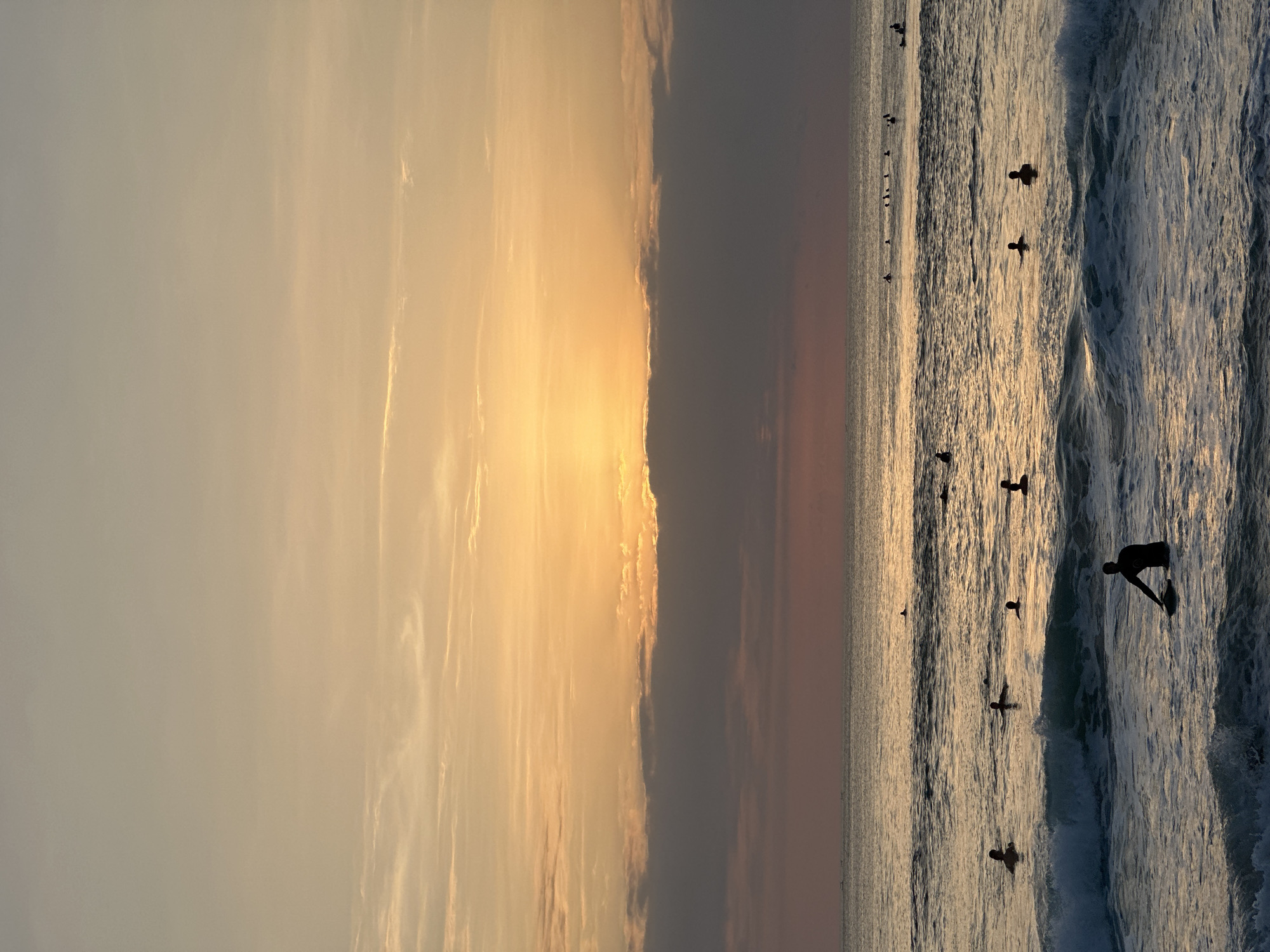 The image shows a group of surfers in the ocean at Kuta Selatan, Indonesia during sunset. The sky is a gradient of orange and yellow, with some clouds obscuring the setting sun. The ocean is calm with gentle waves, and the surfers are scattered across the water, some paddling and others waiting for waves. The overall atmosphere is peaceful and serene.