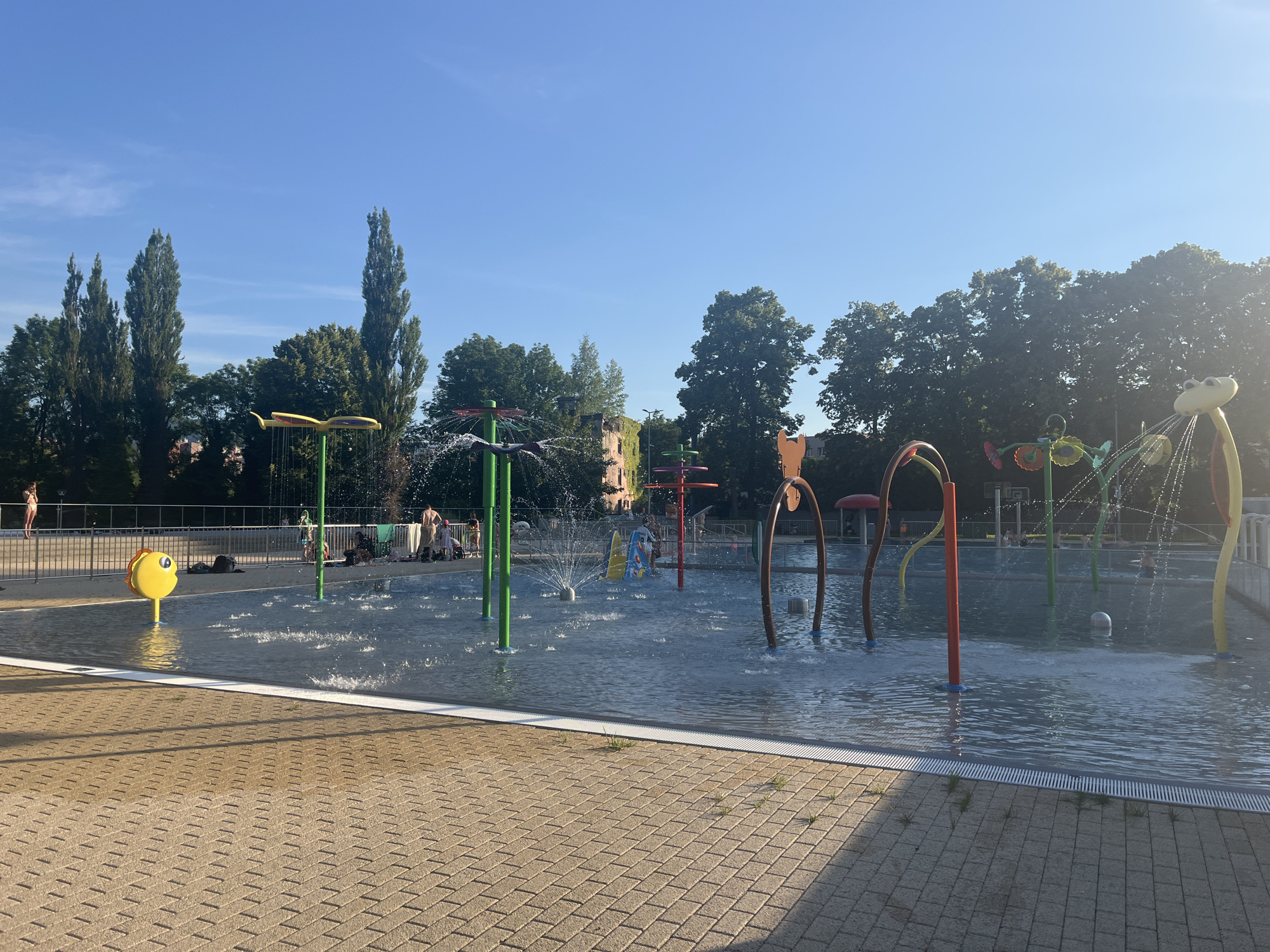 A bright, sunny day at an outdoor water park with various water features. In the foreground, a paved area with a herringbone brick pattern leads to the shallow water of the splash pad. A bright yellow, fish-shaped water dispenser is visible on the left. The water park features several colorful spray fountains and arching water structures, with water actively spraying in several locations. Several children and adults are present, some wading in the water and others sitting around the edges. A metal fence encloses the area, and beyond it, tall trees and some buildings can be seen under a clear blue sky.