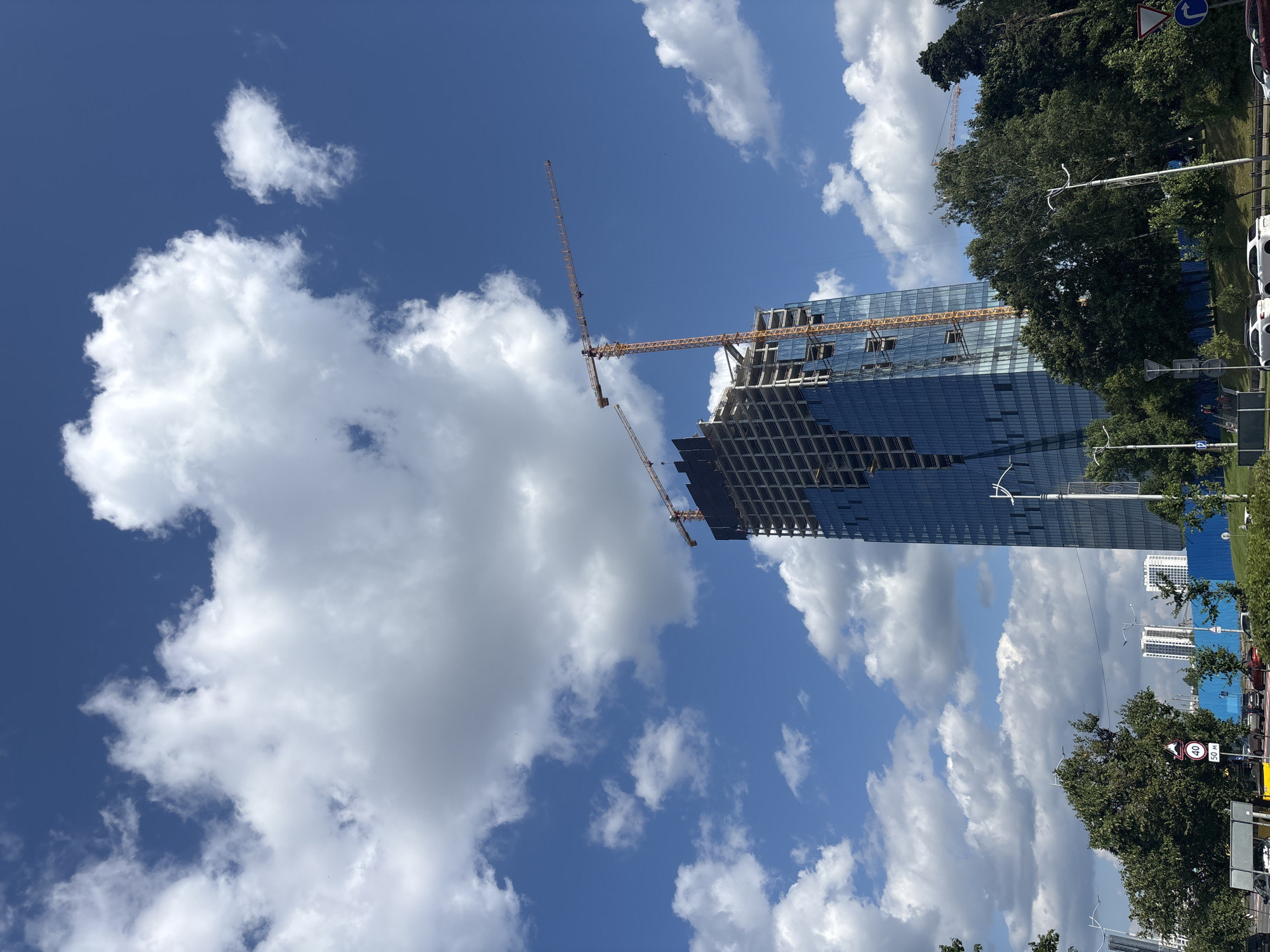 A partially constructed skyscraper with a glass facade and exposed concrete framework dominates the center of the frame. Two yellow construction cranes are visible, one attached to the side of the building and another extending from the top. The sky above is a clear blue, punctuated by fluffy white clouds. In the foreground, a tree-lined street with cars and traffic signs is visible. A blue construction fence runs along the middle ground, with several white apartment buildings in the distance. The image appears to be taken from a street-level perspective, looking up at the skyscraper.