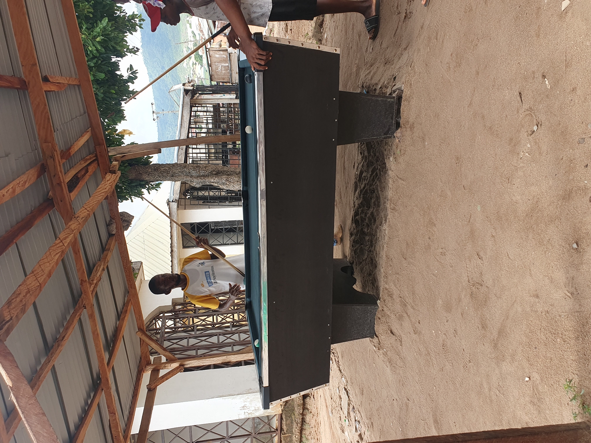 Two men are playing pool on an outdoor billiard table near a beach. One man is wearing a white and yellow shirt and holding a pool cue, aiming at a ball on the table. The other man, wearing a red cap and a patterned shirt, is adjusting the edge of the table. The pool table is positioned on a sandy surface. The background reveals buildings, a metal fence, and a glimpse of the sea with trees and foliage. The location appears to be a beachside setting in Lokoja, Nigeria, characterized by a simple shelter with a wooden frame.