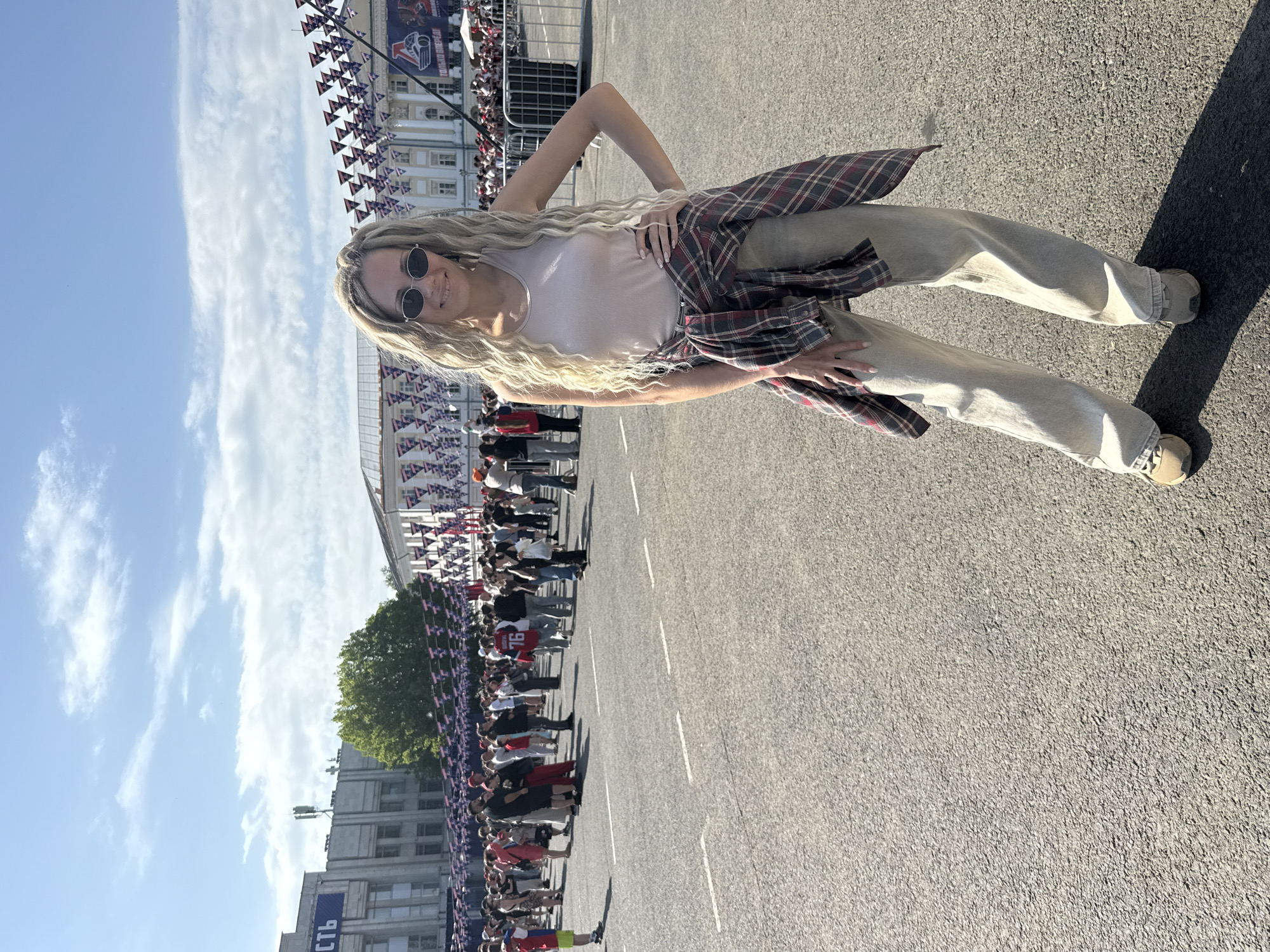 A woman with long blonde hair, sunglasses, and a flannel shirt tied around her waist stands on an asphalt surface. Behind her is a crowd of people lining the street with a building in the background and flags hanging overhead. The location is in the city of Unknown, Unknown.