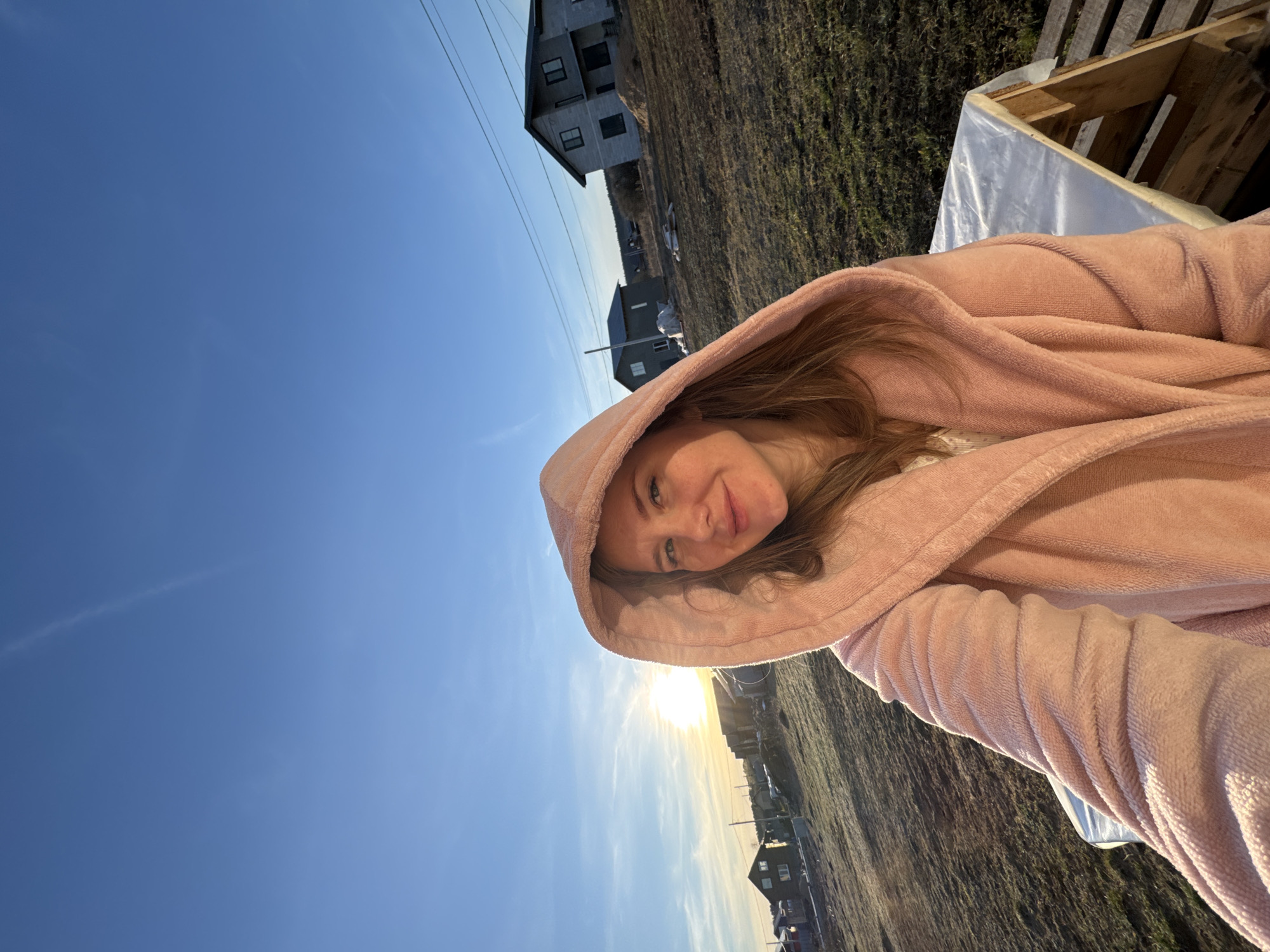 The image features a young woman smiling and looking directly at the camera, her head tilted to the side due to the photo's portrait orientation. She is wearing a soft, fleecy pink hooded robe, with her light brown hair visible around her face, framed by the hood.
The outdoor setting suggests a rural or semi-rural area. The background shows a clear blue sky with the sun low on the horizon, emitting a warm, golden glow, indicating either sunrise or sunset. The ground is covered with dry grass or sparse vegetation. In the middle distance, residential houses with dark roofs and light facades are visible, along with utility poles and wires stretching across the frame. Closer to the foreground, some wooden structures, possibly pallets or crates, partially covered with white plastic sheeting, can be seen.
The woman conveys a cheerful and content emotion through her smile. No visible text is present. This scene is located in Gornyi (Dvurechenskoe S/P), Russia.