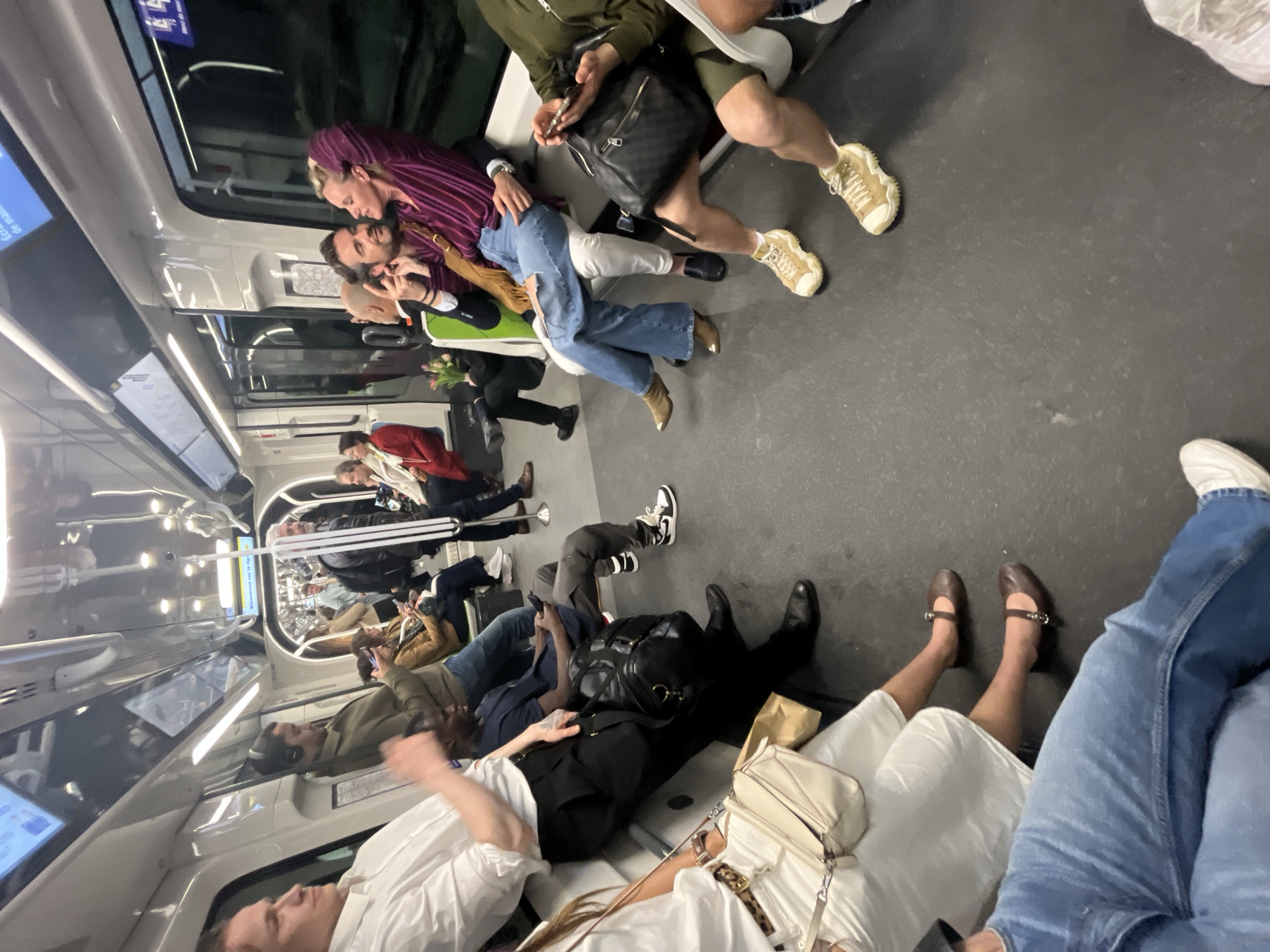 The image captures the interior of a subway car in Paris, France, crowded with passengers. The car's interior features light gray walls and dark gray flooring. Several individuals are seated, while others stand. One couple is embracing near a window. Clothing styles vary among the passengers, with a mix of casual and more formal attire visible. Several bags and personal belongings are also scattered about. The lighting is bright, typical of interior subway car illumination.