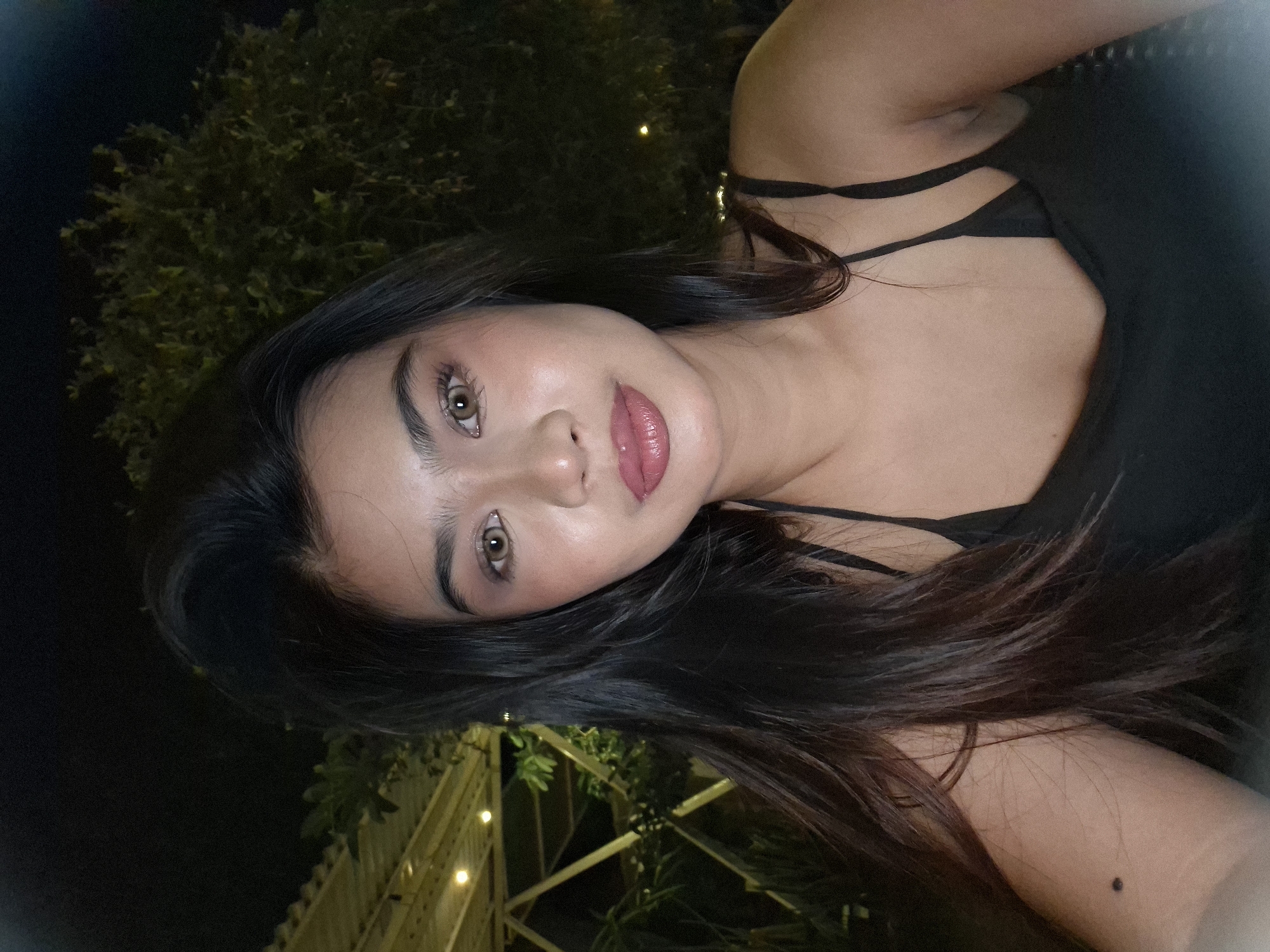 A medium shot photograph captures a young woman with dark hair and light brown eyes, facing the camera at night. She is wearing a black camisole. Her makeup includes foundation, blush, eyeshadow, eyeliner, mascara, and lipstick in a berry shade. The background shows a dimly lit outdoor setting with foliage and structures that appear to be part of a resort or restaurant in Kuta, Indonesia. The lighting suggests artificial light sources, likely from lamps or string lights, are present.