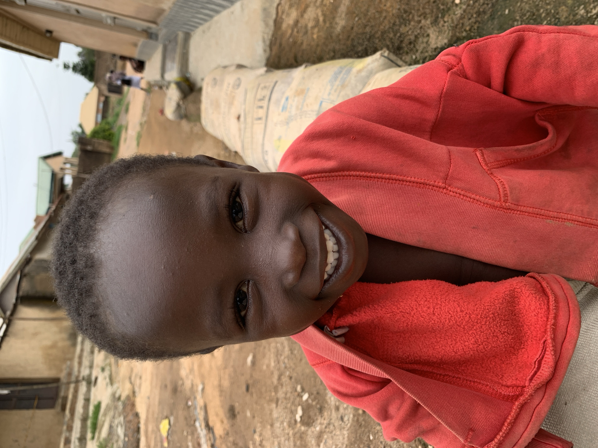 A close-up shot shows a young Black child, appearing to be around 3-5 years old, smiling widely. The child has dark skin and short, dark hair. They are wearing a bright red hooded jacket, with the hood pulled back. The background is blurred, but appears to be an outdoor urban setting, possibly a street or alleyway. The ground is dirt and there are buildings visible in the distance, consistent with an area in Jos, Nigeria. A large, cylindrical object, possibly a sack of cement or similar material, is visible behind the child, wrapped in a beige plastic material. The overall lighting suggests it is daytime.