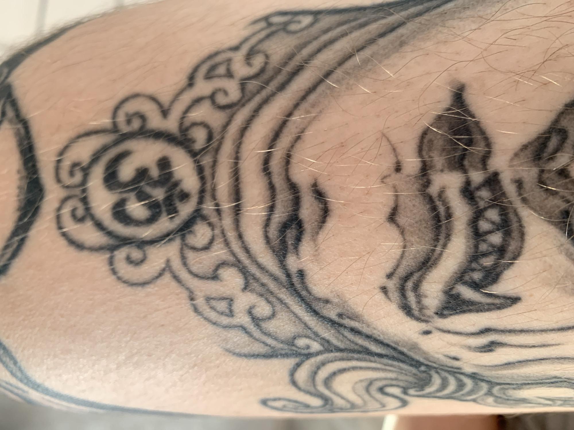 The image shows a close-up of a tattoo on human skin. The tattoo is black and appears to be a decorative design with swirling lines and a central circular motif containing what looks like a symbol or script. The skin has some texture and is covered in light-colored hair. There is no discernible background, indicating the focus is solely on the tattoo itself. The context of Stavropol, Russia, is not visually present in the image.