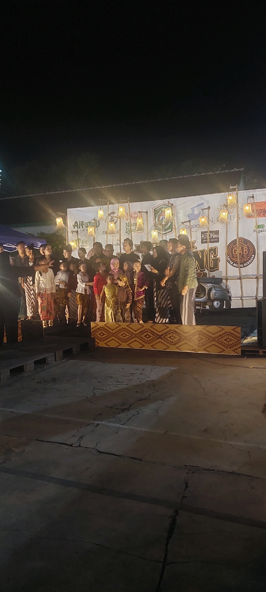 A group of people, including adults and children, are gathered on a stage at night. They appear to be participating in an event, possibly a cultural performance or awards ceremony, given the presence of a backdrop with logos and text. The children are dressed in traditional Indonesian attire, such as batik and kebayas. The stage is decorated with bamboo structures and traditional lamps, illuminated by warm light. In the background, a vintage scooter is visible. The ground in front of the stage is a cracked concrete surface. The event seems to be taking place outdoors, under the night sky. While the specific location in Lumajang, Indonesia, is not explicitly stated in the visual information, the context suggests it is a public event organized within the city.