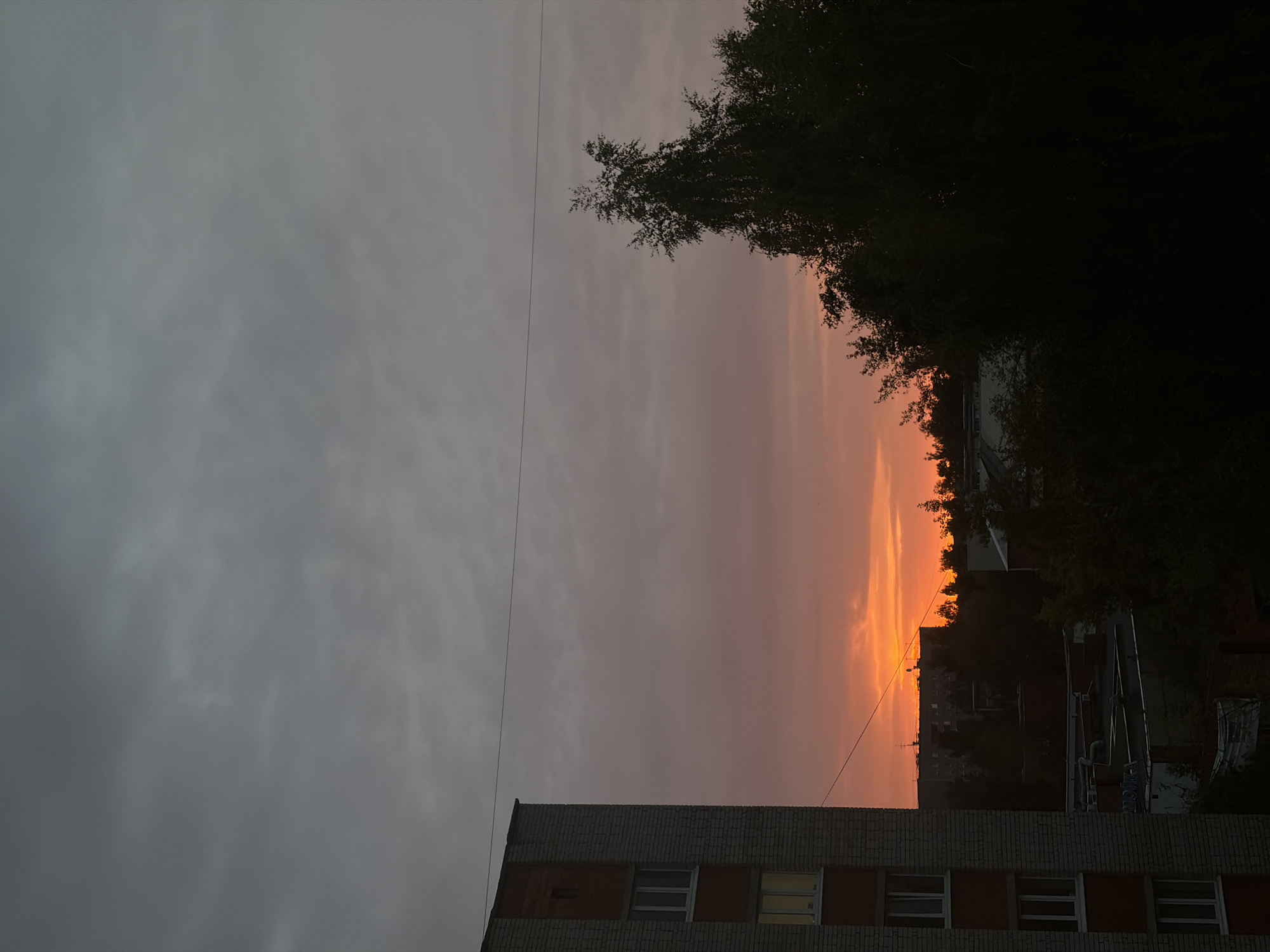This media file captures a dramatic sunset over an urban landscape, oriented vertically. The sky is a striking gradient, transitioning from a muted gray on the left to vibrant shades of orange and yellow near the horizon on the right, indicating it's either sunrise or sunset. Wispy clouds are visible, catching the light of the setting or rising sun.
In the foreground, the top of a brick building is visible, with multiple windows. The building appears to be a multi-story residential or commercial structure. Silhouetted against the colorful sky, a dense line of trees forms a dark, irregular border along the horizon. Beyond the trees, the shapes of other buildings and structures of the city can be vaguely discerned, though they are largely obscured by the trees and the fading light.
The overall mood is one of natural beauty juxtaposed with the built environment. The strong contrast between the dark foreground elements and the bright sky creates a visually impactful scene. The time of day is clearly indicated by the sunset or sunrise colors in the sky. The weather appears to be partly cloudy. No people are visible in the frame. There is no discernible text in the image.