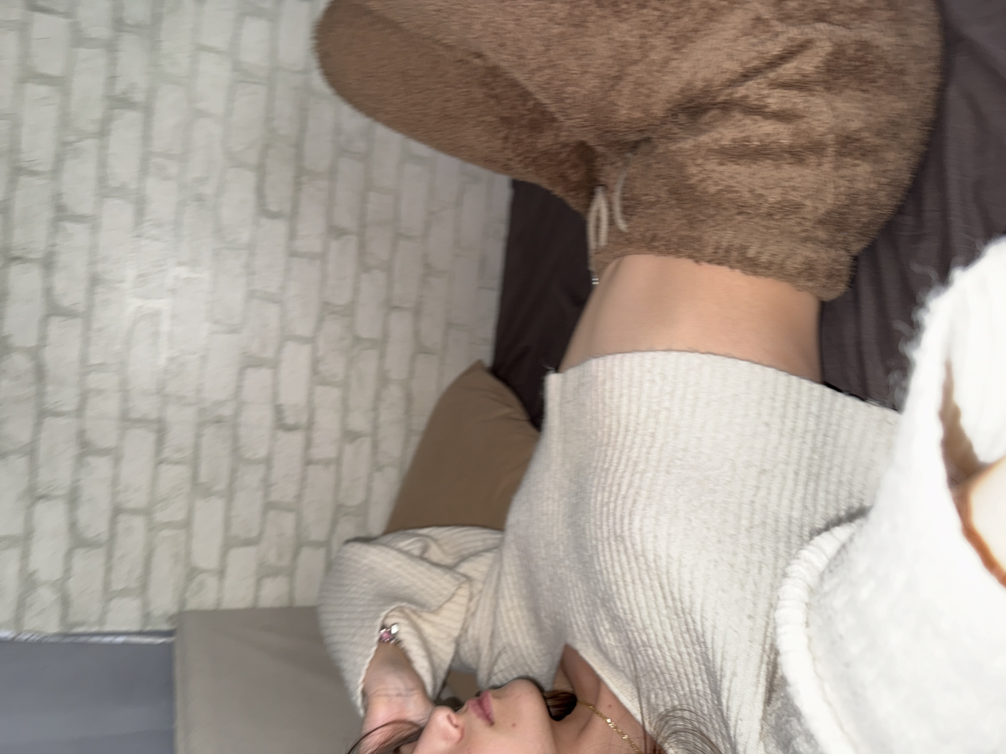 A close-up shot of a person with dark hair, wearing a cream-colored knit sweater and brown sweatpants. The person is lying on a bed or sofa, with a tan pillow nearby. The background includes a grey brick wall. The location is unknown.
