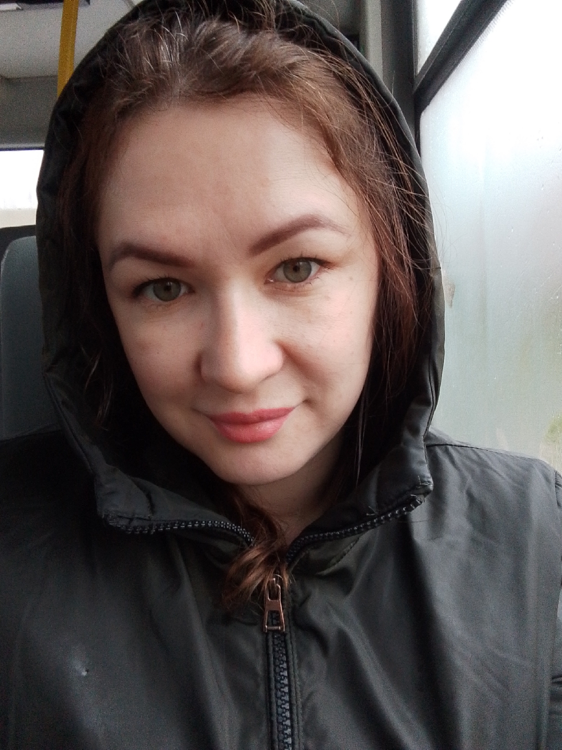 A woman with reddish-brown hair and green-hazel eyes is shown in a close-up, head-and-shoulders portrait. She is wearing a dark, possibly olive green, hooded jacket with the hood up, framing her face. The jacket is zipped up, revealing a bronze-colored zipper pull. She has light makeup, with defined eyebrows and pink lipstick, and exhibits a gentle, subtle smile while looking directly at the viewer.
The setting appears to be inside a public transport vehicle, such as a bus. A window on the right side of the image is covered in condensation or fog, indicating cold or humid weather outside. A faint yellow bar or handle from the vehicle's interior is visible in the upper left background. The overall lighting is soft, suggesting either an overcast day or an interior setting with muted light. The scene is consistent with travel in a potentially chilly environment, given the location context of Chaikovsky, Russia. There is no visible text.