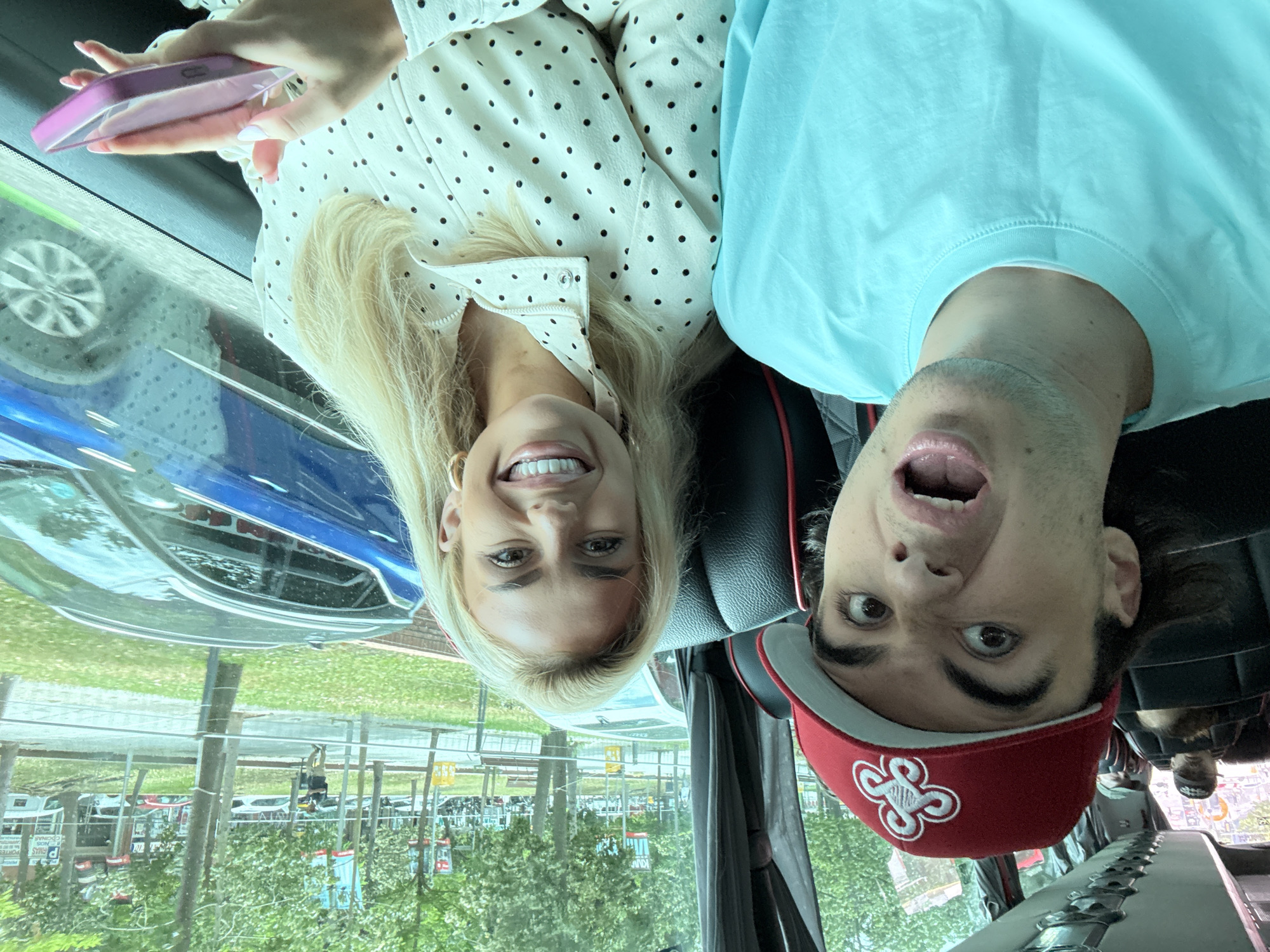 The image depicts a woman and a man taking a photo inside a vehicle, likely a car or bus, with the image rotated 90 degrees clockwise, making the subjects appear upside down.
The woman, positioned on the left, has long blonde hair and a bright smile. She is wearing a white long-sleeved shirt adorned with black polka dots. She holds a pink smartphone in her left hand, which has light-colored nail polish.
To her right, the man wears a light blue t-shirt and a red baseball cap with a distinctive white, stylized clover-like logo. He has dark hair, and his face shows a playful or surprised expression with wide-open eyes and an open mouth.
Through the vehicle's window, an outdoor scene is visible, illuminated by daylight. Lush green trees and grass are prominent, alongside what appears to be a parking area or street with several other vehicles. Reflections of the exterior, including a blue car, are noticeable on the window. In the distant background, some blurry signs with faint text are discernible; one sign partially displays "P LUS" and "COCINA."
The overall atmosphere is cheerful, conveyed by the subjects' expressions. The scene is set during the daytime, and the location context is Barcelona, Spain.