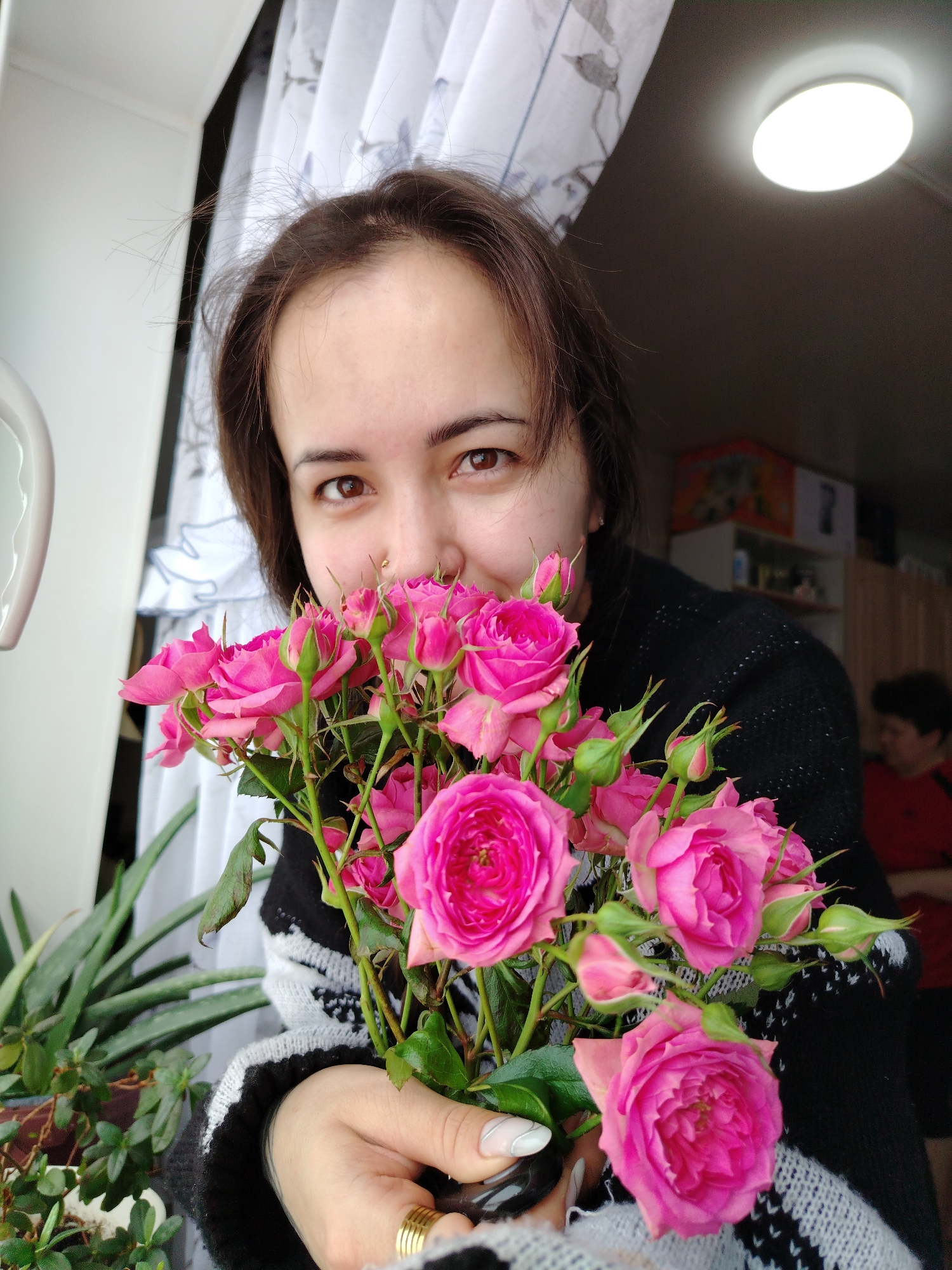 An indoor scene in Гожан, Russia, depicts a woman in her 20s-30s holding a bouquet of pink roses. She has dark brown hair, brown eyes, and a small gold stud piercing on her right nostril. She is wearing a dark sweater with a white/grey patterned design on the sleeves. Her left hand, adorned with a gold multi-band ring on the ring finger and white-tipped fingernails, supports the roses which partially obscure her lower face. She smiles faintly, looking towards the camera. In the background to the left, a white wall or window frame is visible, alongside white curtains with a subtle dark pattern, and two potted green plants. The upper right background shows a dark ceiling with an illuminated circular white light fixture. Further back, out of focus, shelves or cabinets containing various objects are discernible, and a second, blurred individual with short dark hair, wearing a red garment, is partially visible.