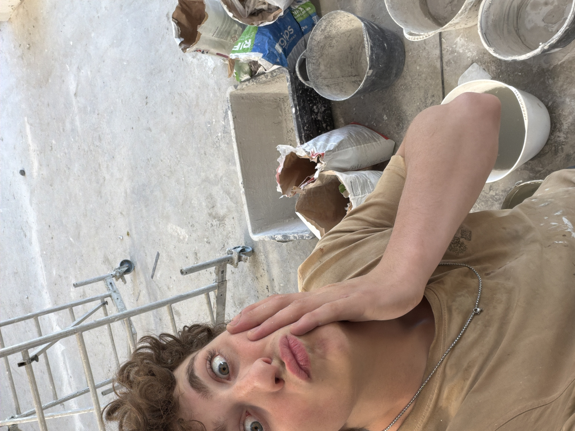 The image shows a person with curly brown hair and a surprised expression, with one hand on their cheek. They are wearing a tan t-shirt and a silver necklace. The person appears to be in an unfinished building or construction site, with a concrete floor and scaffolding visible in the background. There are bags of what appear to be construction materials, such as plaster or cement, scattered around, as well as several buckets. One bucket contains a gray liquid, and the person's hand is submerged in it, suggesting they are mixing materials. The overall scene depicts a moment of playful surprise or mock disbelief amidst a work-in-progress environment.