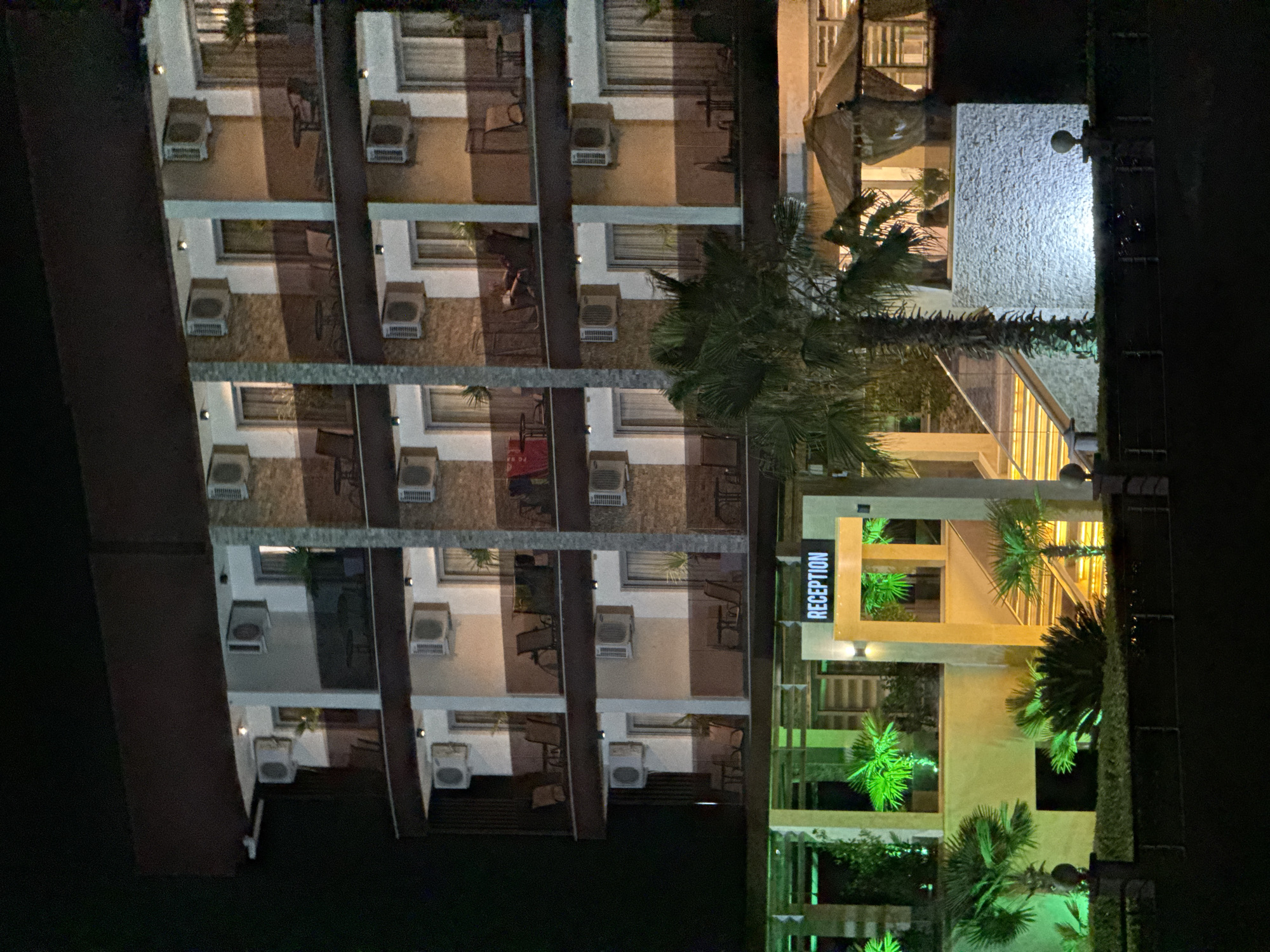 The image captures a nighttime view of a modern hotel in Donji Štoj - Shtoji i Poshtëm, Montenegro. The hotel features multiple floors with balconies, each equipped with air conditioning units and railings. Balconies have chairs and tables, and some rooms have curtains visible. The hotel's facade is a mix of white, brown, and stone-tiled sections. A prominent "RECEPTION" sign is illuminated above an entrance area with stairs. The foreground is landscaped with palm trees and illuminated foliage in green and yellow, creating a vibrant ambiance. A covered seating area with draped curtains is visible to the right. The scene is set against a dark, night sky, with no people clearly discernible.
