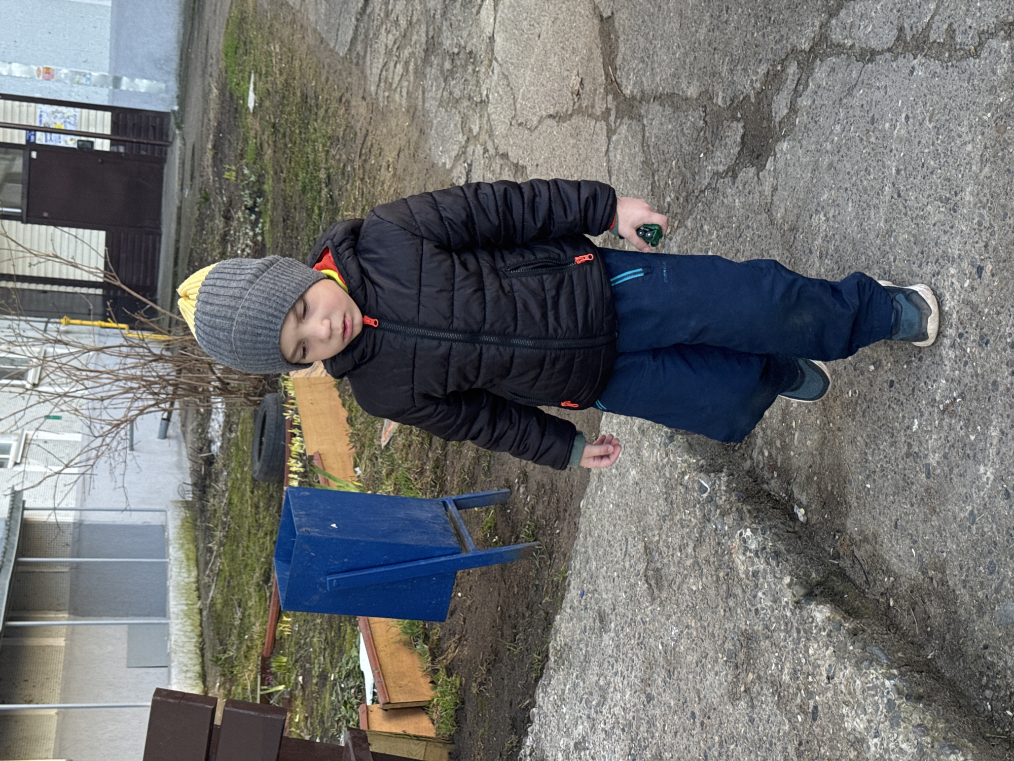 A young boy stands outdoors, centrally positioned in the frame, wearing a black puffer jacket with orange zipper details, a grey beanie with a yellow stripe, navy blue pants, and dark sneakers. He holds a small, dark green object, possibly a toy, in his right hand. His expression is neutral, and he appears to be looking slightly downwards or to his left.
The setting is an outdoor, somewhat utilitarian area, likely a residential courtyard or back alley in an urban environment. The ground beneath him is cracked asphalt and concrete, transitioning to patches of dirt and sparse green grass. In the background, to his left, are bare trees and a light-colored multi-story building with a dark brown entrance door and windows. To his right, closer to the foreground, is a bright blue, open-top trash bin, a discarded car tire, and some wooden planks.
The time of day is daytime, but the light is soft, suggesting an overcast sky or perhaps early morning/late afternoon. The boy's warm clothing indicates cool weather. The environment appears lived-in but also somewhat neglected, typical of older residential areas. No legible text is visible on any objects or surfaces within the image. The overall atmosphere is quiet and mundane.