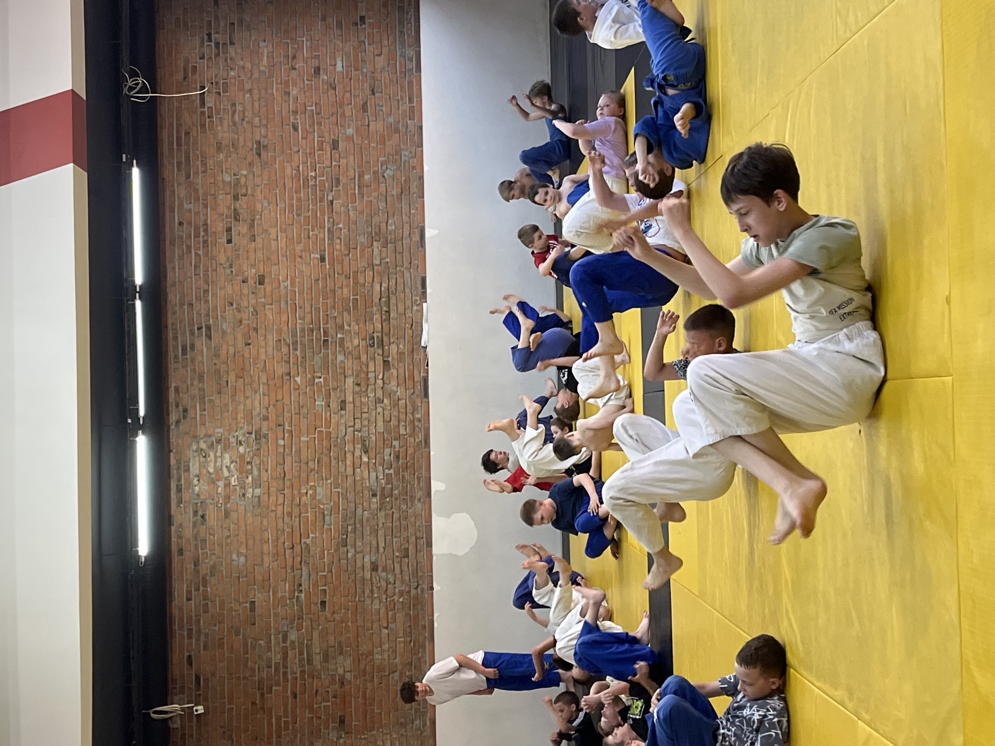 A photograph depicts a large group of children in what appears to be a judo class in Krasnoyarsk, Russia. They are positioned on a yellow mat, some sitting and others seemingly practicing moves. Most of the children are wearing judo uniforms, a combination of white and blue. A teenage boy in a white judo outfit is sitting in the foreground. In the background is a wall made of brick, and above the group, fluorescent lights are mounted on the wall.