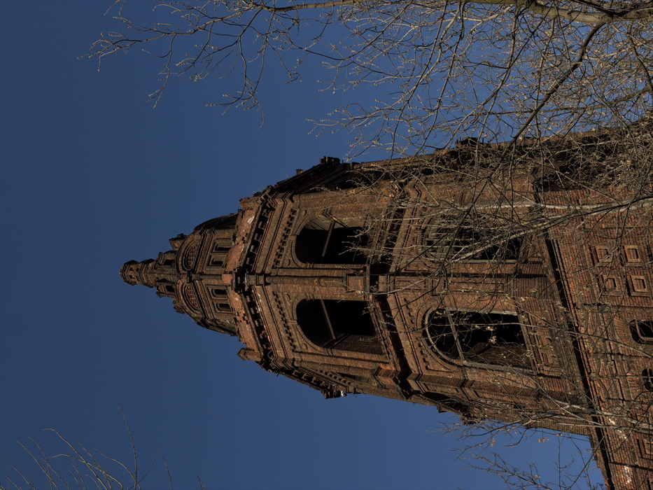 A low-angle photograph captures the imposing, multi-tiered bell tower of the Rogozhsky Old Believer Community Cultural and Business Center, located in Moscow, Russia, against a clear, deep blue sky. The structure is constructed from dark red brick, exhibiting intricate masonry work, decorative cornices, and multiple arched and rectangular apertures on its upper levels. These openings are vacant, lacking windowpanes or shutters, revealing darkened interiors. The tower culminates in a tapering spire, also crafted from brick, with small, decorative arched openings. Numerous bare, leafless deciduous tree branches, some displaying visible buds, partially frame and overlap the tower on both sides and in the foreground. Direct sunlight illuminates the tower from the viewer's left, highlighting the weathered brick texture. No individuals or animated subjects are present in the scene.