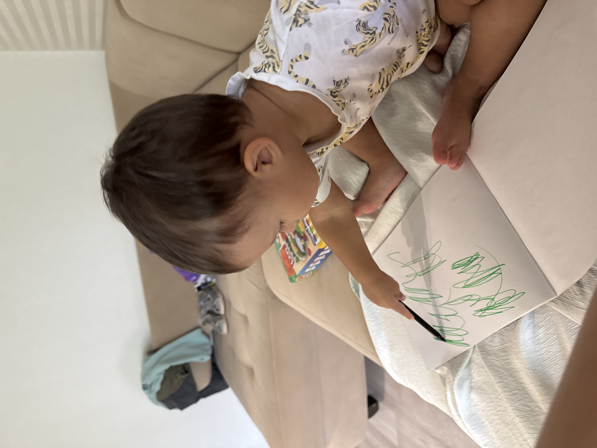 An indoor, high-angle shot captures a young child, appearing to be a toddler, with dark hair, focused on drawing. The child, wearing a white short-sleeved top adorned with yellow and black tiger patterns, holds a black marker in their right hand, producing green scribbles on a white open book or pad. The child is partially positioned on a light-colored, textured fabric surface. In the background, a light beige upholstered sofa is visible, holding various items: a folded teal garment and dark fabric on the left, a pair of light-colored athletic shoes centrally, and a colorful box, possibly a toy or game, with red, green, and blue details, to the right. A plain white wall forms the far background, with a section of vertical blinds or textured wall visible on the right. A light-colored floor is visible in the lower-left corner. The visual content of the image does not contain any identifying elements, landmarks, or text that would indicate the specific location as Chaikovsky, Russia.