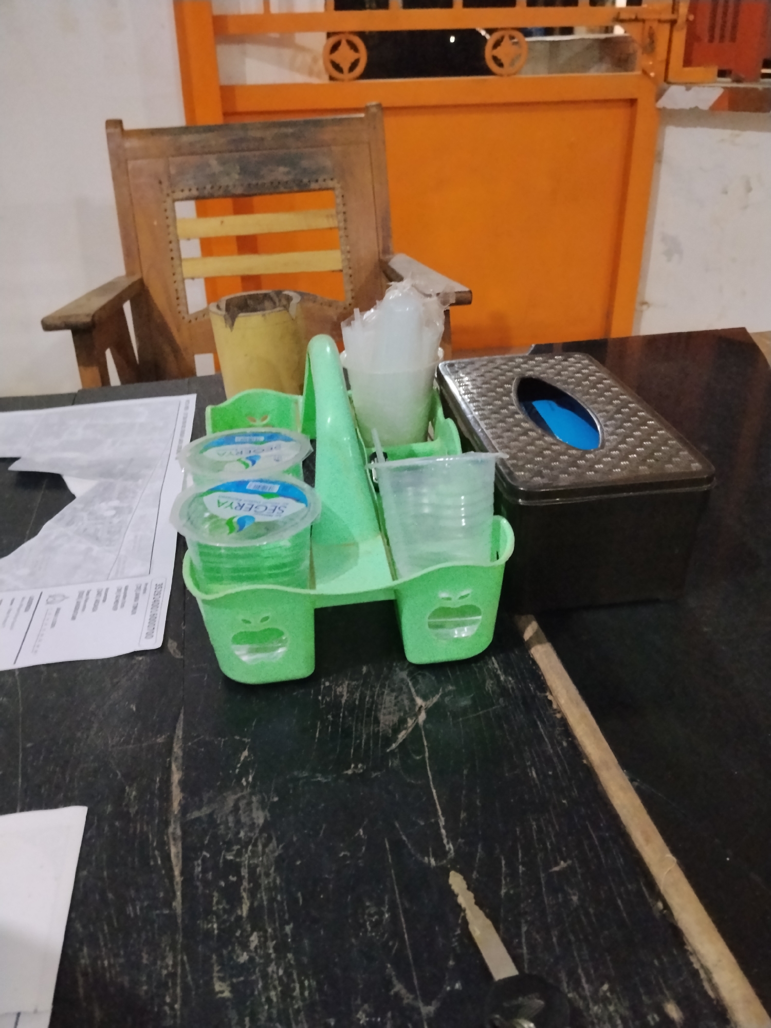 The image displays a tabletop with various items, including a green plastic cup holder containing several plastic cups of water, a black tissue box, and some papers. In the background, there is an orange structure and a wooden chair. The scene appears to be indoors, possibly in a waiting area or a dining establishment. There are no people visible in the image, and no actions or interactions are depicted. The lighting suggests it might be evening or a dimly lit interior.