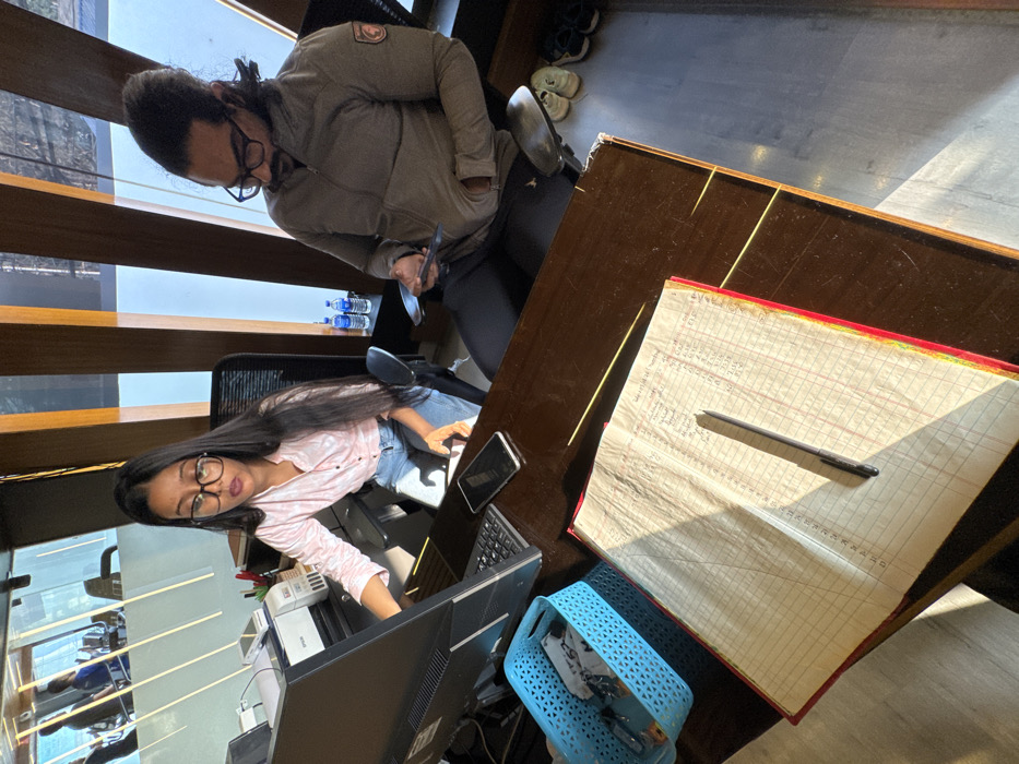 Two individuals are in an office setting. One person, wearing glasses and a light pink shirt, sits in front of a computer and monitor, with a blue basket nearby. The second individual, also wearing glasses and a gray jacket, stands behind the first person, holding a phone. A large, aged ledger with handwritten notes and a pen rests on a desk. Several water bottles are visible near the window blinds. Additional shoes are near the corner of the room.