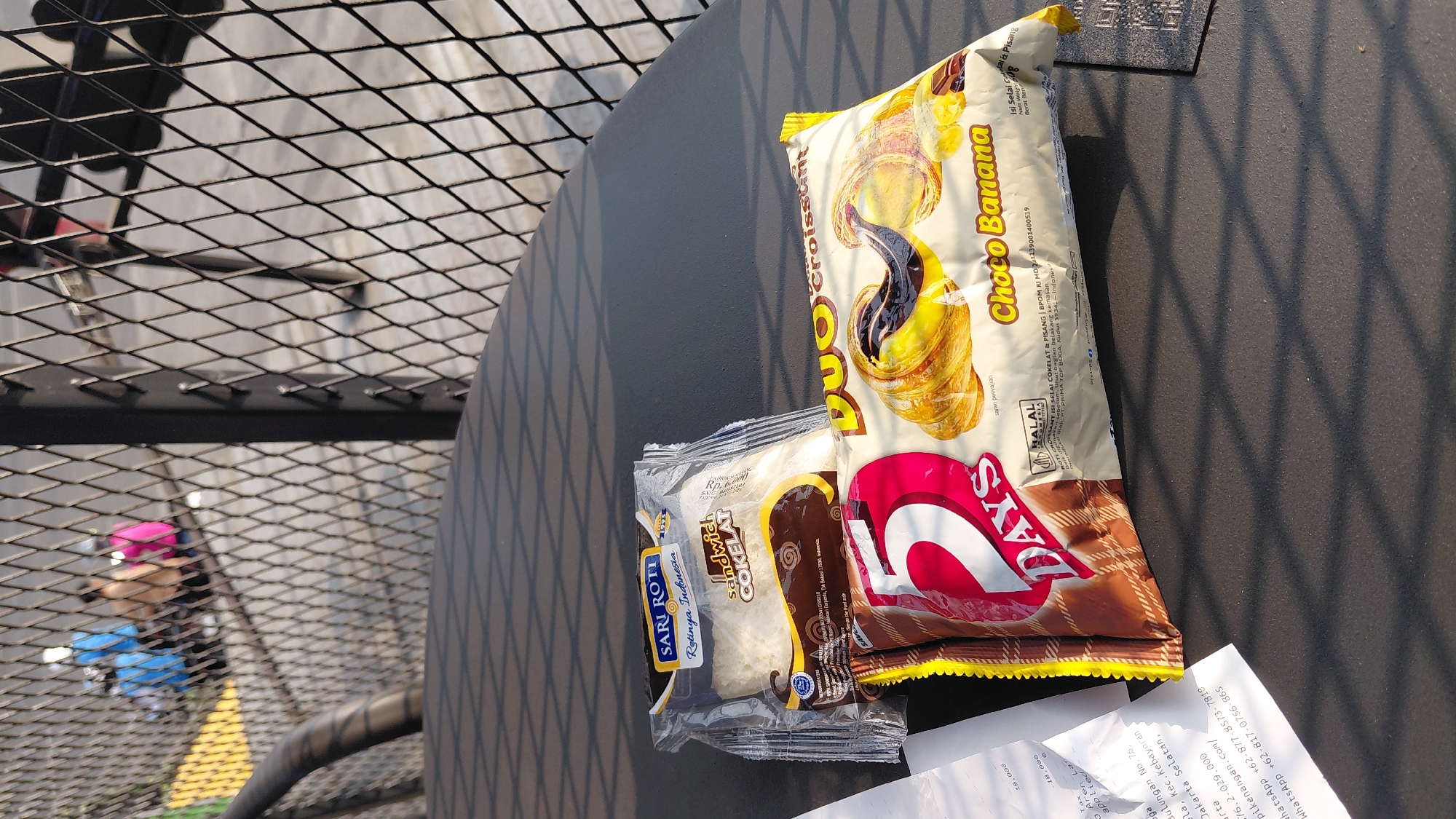 In Karanglewas, Indonesia, two snack packages are placed on a dark, textured surface. The package on the left is transparent, revealing white bread labeled "Sari Roti Rotiya Indonesia Sandwich Cokelat". The package on the right is yellow and features an image of a croissant with chocolate filling and banana slices, labeled "Duo Croissant Choco Banana". A white receipt, partially visible, is positioned to the left of the snack packages, displaying text in Indonesian. In the background, a black metal mesh fence creates a grid pattern, with blurry figures of people visible beyond it, suggesting an outdoor or semi-outdoor setting. The lighting indicates it is daytime, casting shadows on the surface.