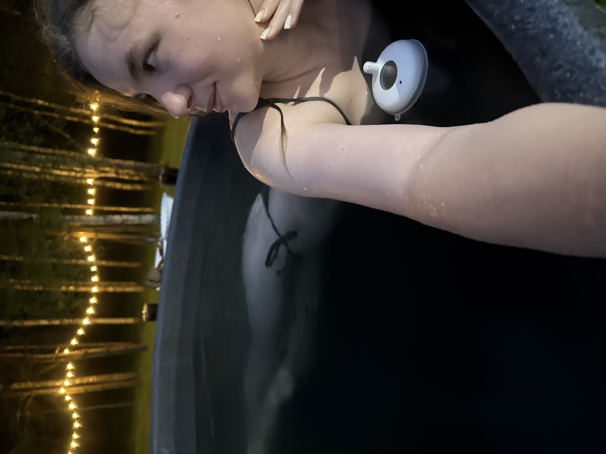 The image depicts an outdoor nighttime scene in Копнино, Russia, featuring a young adult female in a dark-colored tub or plunge pool. The woman is positioned with her right shoulder and head facing the right side of the frame, gazing upward and to the right. She has fair skin, dark hair with visible wet strands, and water droplets are discernible on her face, neck, and left arm. She is wearing a dark-colored strap top. Her left arm is raised and partially visible in the foreground. Her right hand, displaying light-colored fingernail polish, is near her neck. A white, lidded drink container is partially submerged in the water beside her right side. In the background, a horizontal string of warm, glowing lights illuminates several tall, slender trees with light-colored bark. The water in the tub is dark, and the overall environment is dimly lit, indicating evening or night conditions.