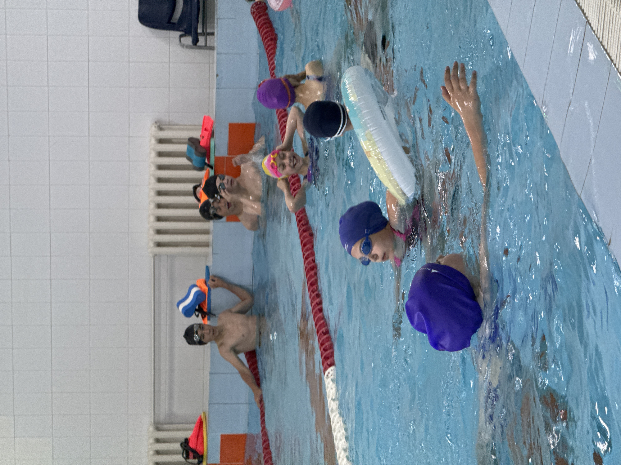 A group of children are participating in a swimming lesson in an indoor pool. The pool is lined with blue tiles and has red and white lane markers. Several children are visible in the water, wearing swim caps and goggles. One boy, appearing to be a teenager, stands in the shallow end, leaning on the pool edge. Younger children are also in the water, some with flotation devices. In the background, there are radiators and a chair. The overall scene depicts a typical swimming lesson for children. The location of Birobidzhan, Russia is not discernible from the image itself.
