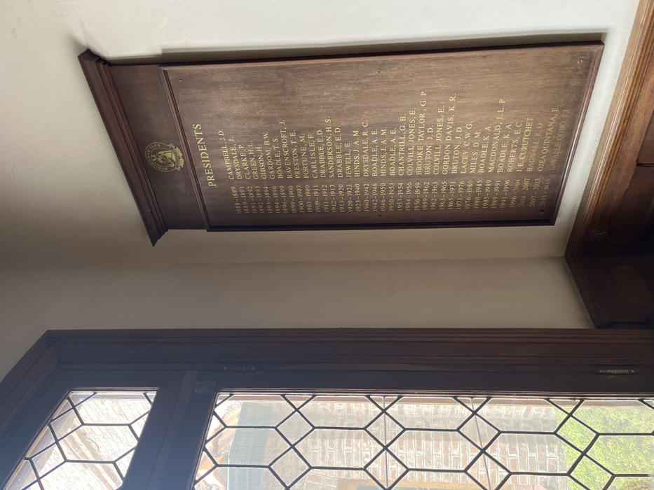 The image displays a long, dark wooden plaque mounted on a light-colored wall, with a leaded glass window visible below it. The image is oriented with the plaque appearing on its side, but the text indicates it's meant to be read horizontally.
The plaque itself is made of a rich, dark brown wood, possibly mahogany or oak, with a slightly glossy finish. It has a decorative molded edge along its top and bottom. On the left side of the plaque, there is a circular golden emblem or crest, though the details of the design are difficult to discern clearly. To the right of the emblem, the word "PRESIDENTS" is inscribed in large, golden, sans-serif capital letters.
Below "PRESIDENTS," a detailed list of names, each accompanied by a date range, is inscribed in smaller, golden capital letters, formatted in two columns. The list spans from 1888-1889 up to 2013-2015. The entries are:
* 1888-1889 CAMPBELL, J. D.
* 1889-1891 DRYSDALE, J.
* 1891-1892 CLARKE, P.
* 1892-1893 GREEN, H.L.
* 1893-1894 GIBSON, H.
* 1894-1896 GARDOM, B.W.
* 1896-1899 BOADLE, T.S.
* 1899-1900 RAVENSCROFT, J.
* 1900-1903 PRESTON, T.E.
* 1903-1909 FORTUNE, M.
* 1909-1911 CARLISLE, F.
* 1911-1912 DRABBLE, E.D.
* 1912-1913 SANDERSON, H. S.
* 1913-1920 DRABBLE, E. D.
* 1920-1923 JEWELL, E.
* 1923-1940 HINDS, J. A. M.
* 1940-1942 DRYSDALE, R. C.
* 1942-1946 BOADLE, A. E.
* 1946-1950 HINDS, J. A. M.
* 1950-1953 BOADLE, A. E.
* 1953-1954 CHANTRILL, G. B.
* 1954-1956 COLVILL JONES, E.
* 1956-1959 BROOKE TAYLOR, G. P.
* 1959-1962 BRUTON, J. D.
* 1962-1963 COLVILL JONES, E.
* 1963-1965 GORDON DAVIS, K. R.
* 1965-1971 BRUTON, J. D.
* 1971-1976 LACEY, C.W.G.
* 1976-1980 MILES, D. M.
* 1980-1985 BOADLE, K. A.
* 1985-1989 MACDONALD, I. L. P
* 1989-1991 BOADLE, K. A.
* 1991-1996 ROBERTS, E.C.
* 1996-2004 J. E. CURUTCHET
* 2004-2007 BAGNALL EO
* 2007-2013 QJEA QUINTANA, E
* 2013-2015 LAVALLE COBO, FJ
The wall behind the plaque is painted a plain, light cream or off-white color. Below the plaque, a window with a dark wooden frame is visible. The window features multiple small panes of glass separated by dark lead cames, forming a hexagonal grid pattern. Through the window, blurred green foliage and what appears to be a light-colored brick structure can be seen, suggesting an outdoor view.
The lighting in the image appears to be natural, coming from the window, casting a soft, even light on the plaque and wall. The photo is taken from a slightly low and angled perspective, looking up towards the plaque and across towards the window.
