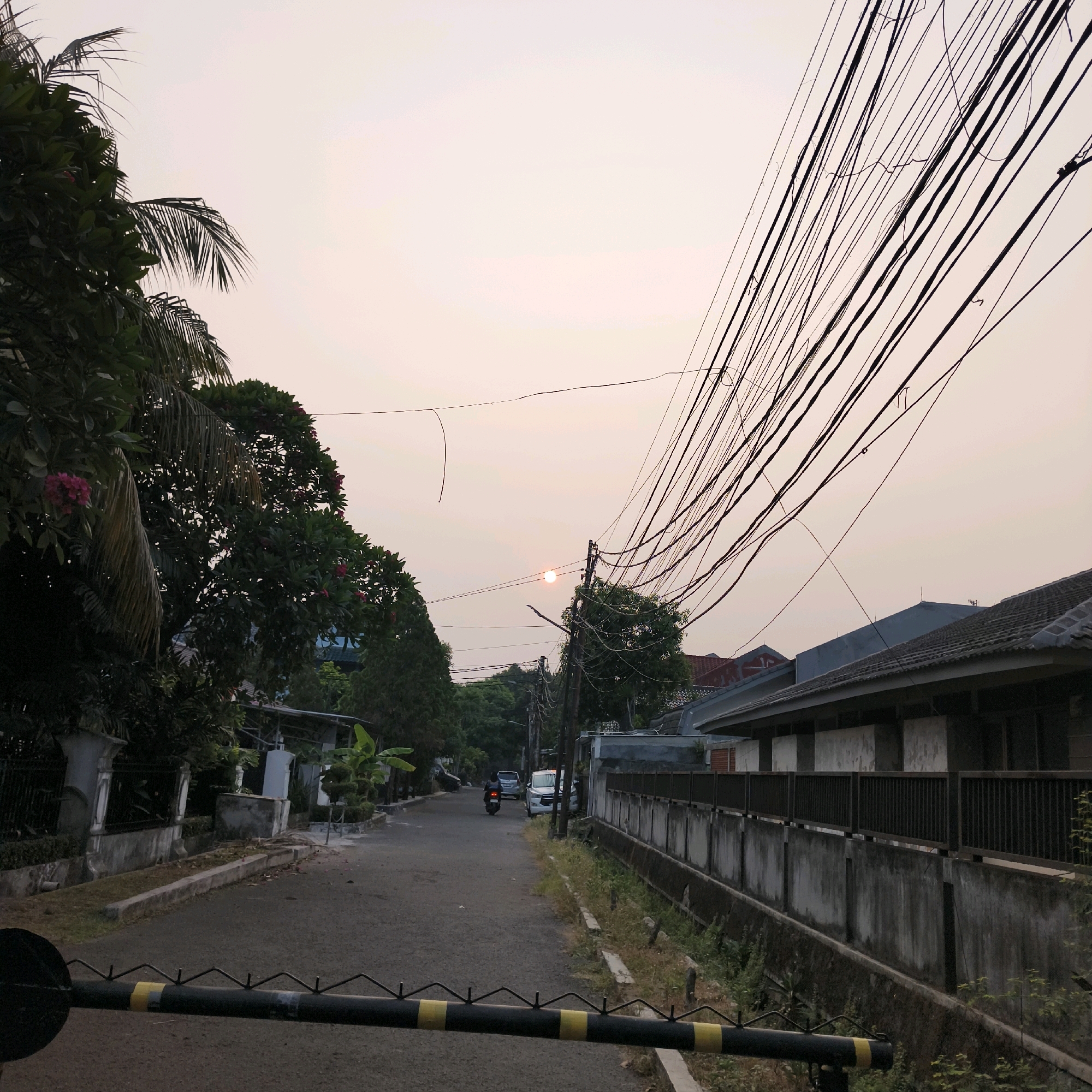 The image depicts a street scene at dusk in South Jakarta, Indonesia. The sky is a pale orange, indicating the sun is low on the horizon, partially obscured by trees and overhead wires. The street is paved, with a single motorcycle and a car visible in the distance. To the left of the street, there are trees and fences surrounding residential properties. To the right, a concrete wall with a metal railing runs along the property line, adjacent to a drainage ditch. A black and yellow striped barrier is in the foreground, suggesting a checkpoint or entrance. The overall impression is that of a quiet urban residential area during twilight.