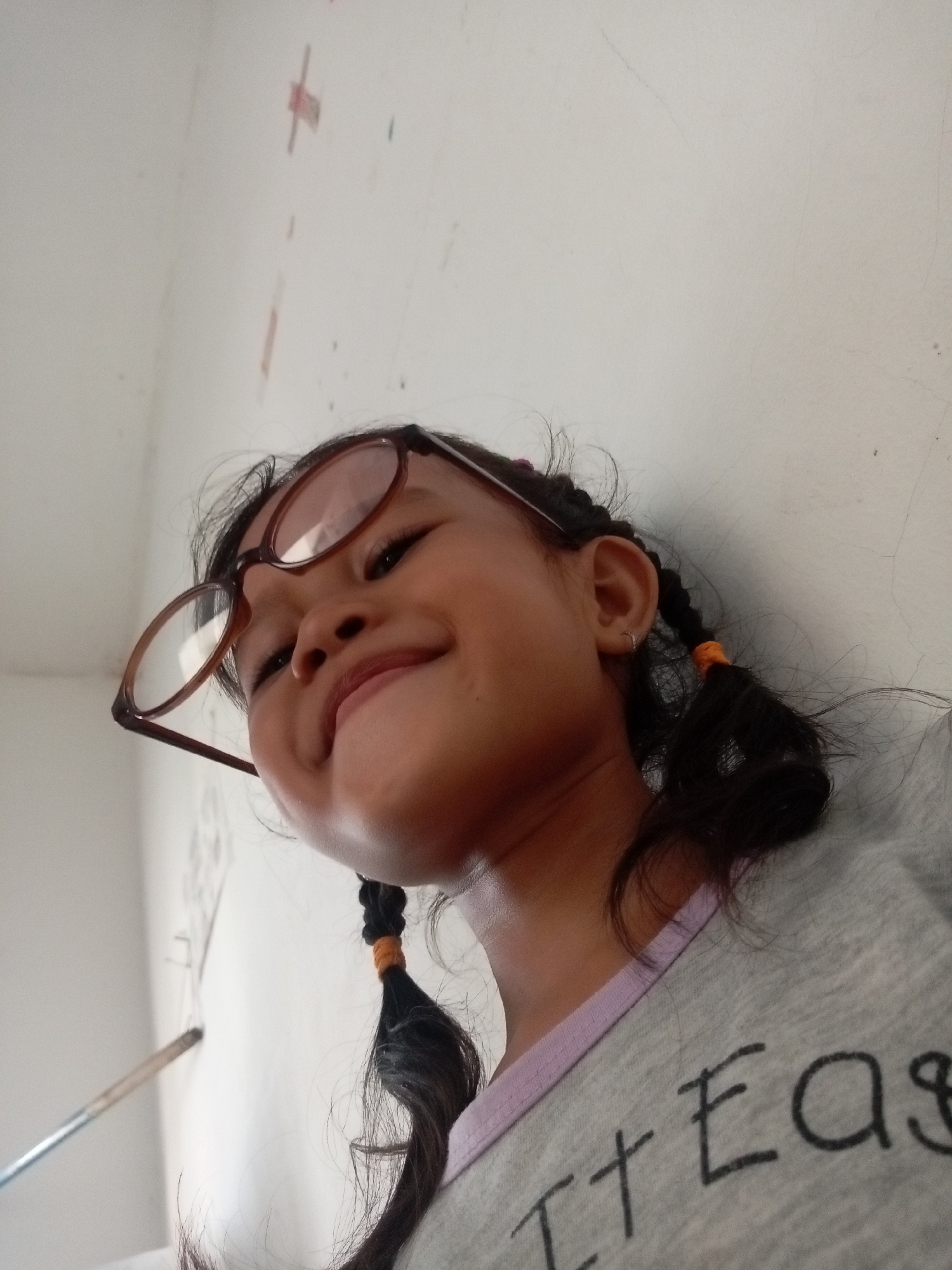 In Batam, Indonesia, a low-angle, close-up, outdoor, portrait-style photograph shows a young girl with dark hair styled into two braids, each tied with an orange band, wearing brown-rimmed glasses. The girl is looking up and to her left, with her mouth slightly open and a subtle smile, her head tilted against a light-colored wall. She is wearing a grey t-shirt with a light purple collar, and on the shirt, partially visible text reads "It Eas". The background is a plain, textured, light-colored wall with faint smudges and what appear to be red marks, possibly from crayons or paint. A silver-colored stick-like object, possibly a paintbrush or pencil, is visible in the lower left corner of the frame. The lighting suggests it is daytime, with shadows indicating light coming from above and slightly to the left.